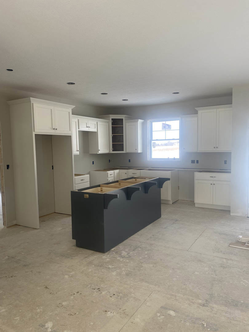White kitchen cabinets with silver knobs, black island with matching countertop, drawers beneath counter, window with sign, light walls and ceiling, hardwood floor.