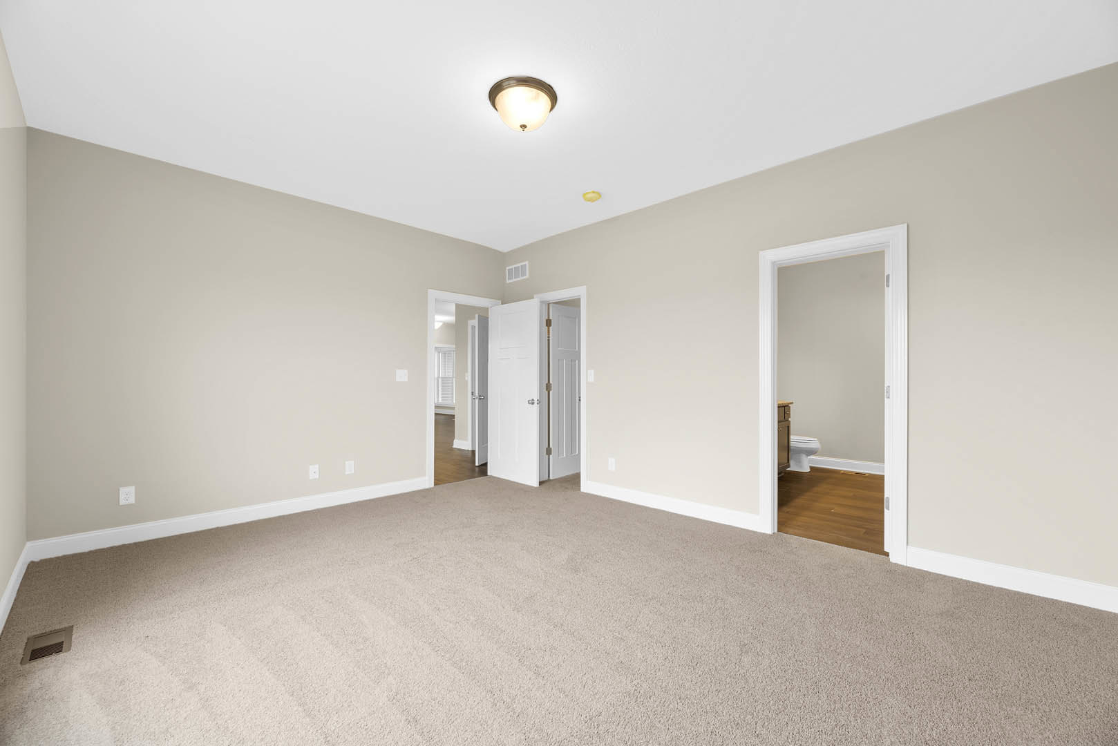 Beige carpeted floor, white walls, ceiling light fixture, white toilet with closed seat, bathroom sink, door with metal bar