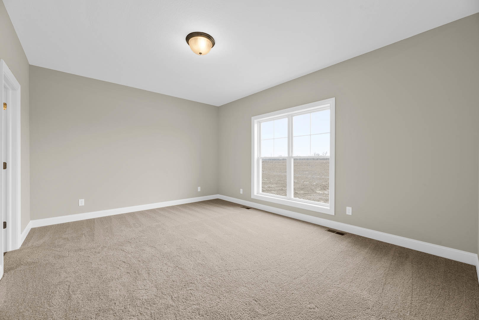 Carpeted room with white walls, large window overlooking grassy field, ceiling light fixture illuminated, door handle visible in foreground, wood trim and molding accents