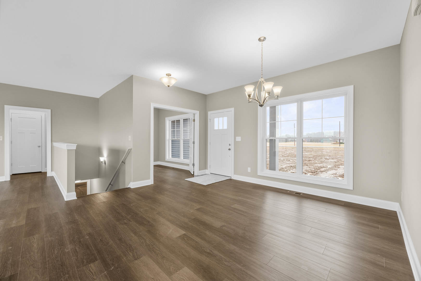 Room with hardwood flooring, white walls, a white door with a silver handle, large window overlooking a dirt field, and a decorative chandelier hanging from the ceiling