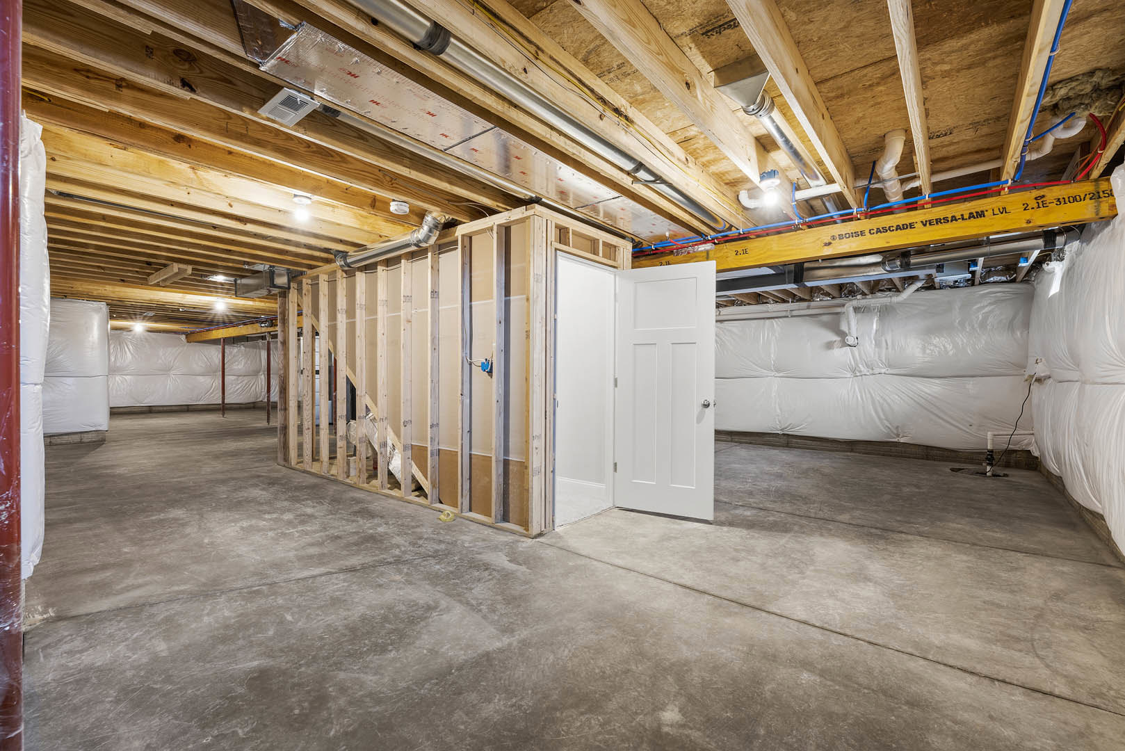Concrete floor and unfinished walls with exposed steel beams and pipes, white door with silver knobs, white plastic bag hanging on wall, basement interior with visible building