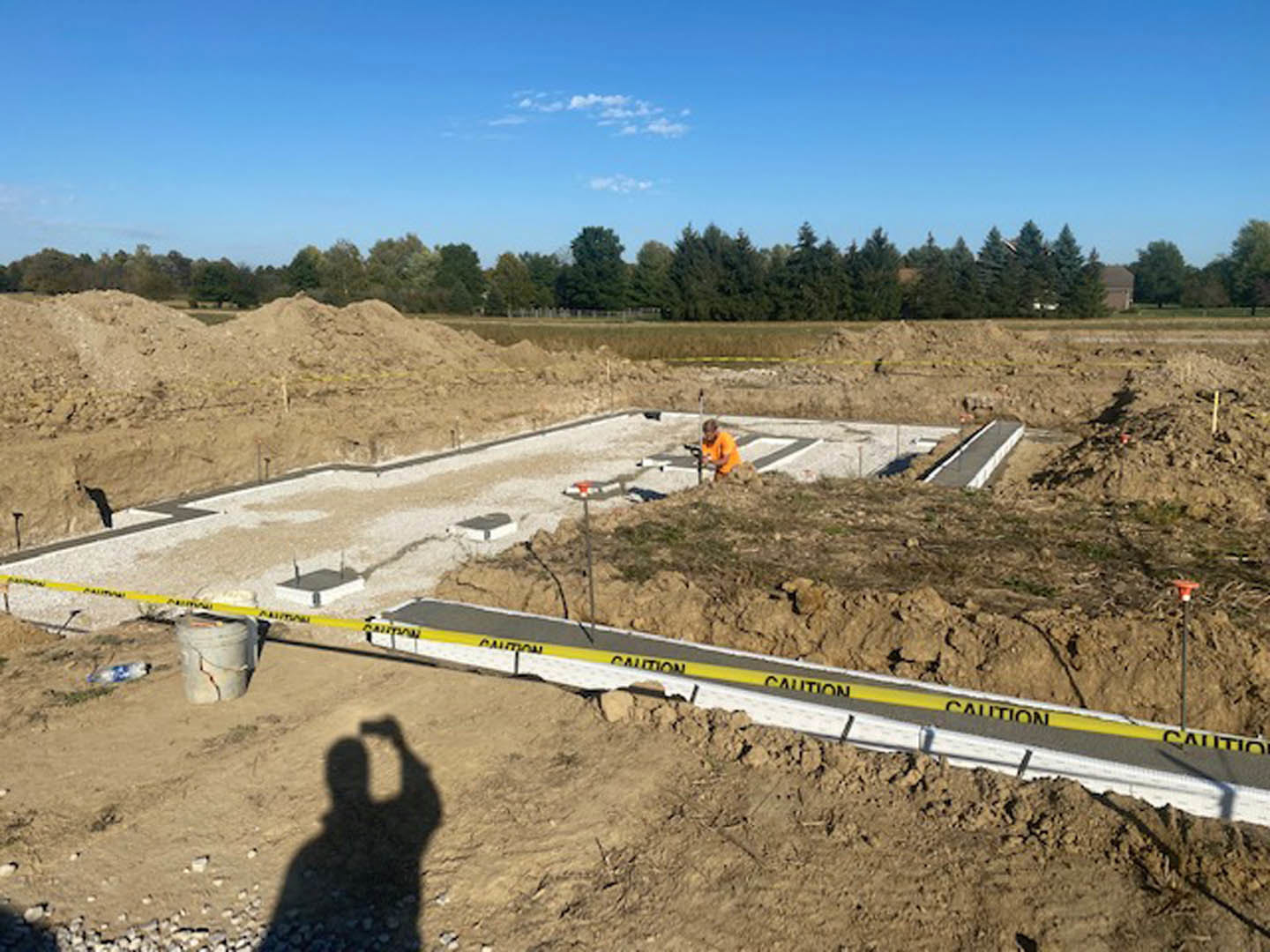 Man in orange shirt working in dirt area near yellow caution tape, with blue sky and scattered clouds overhead, shadow of person taking selfie visible on ground