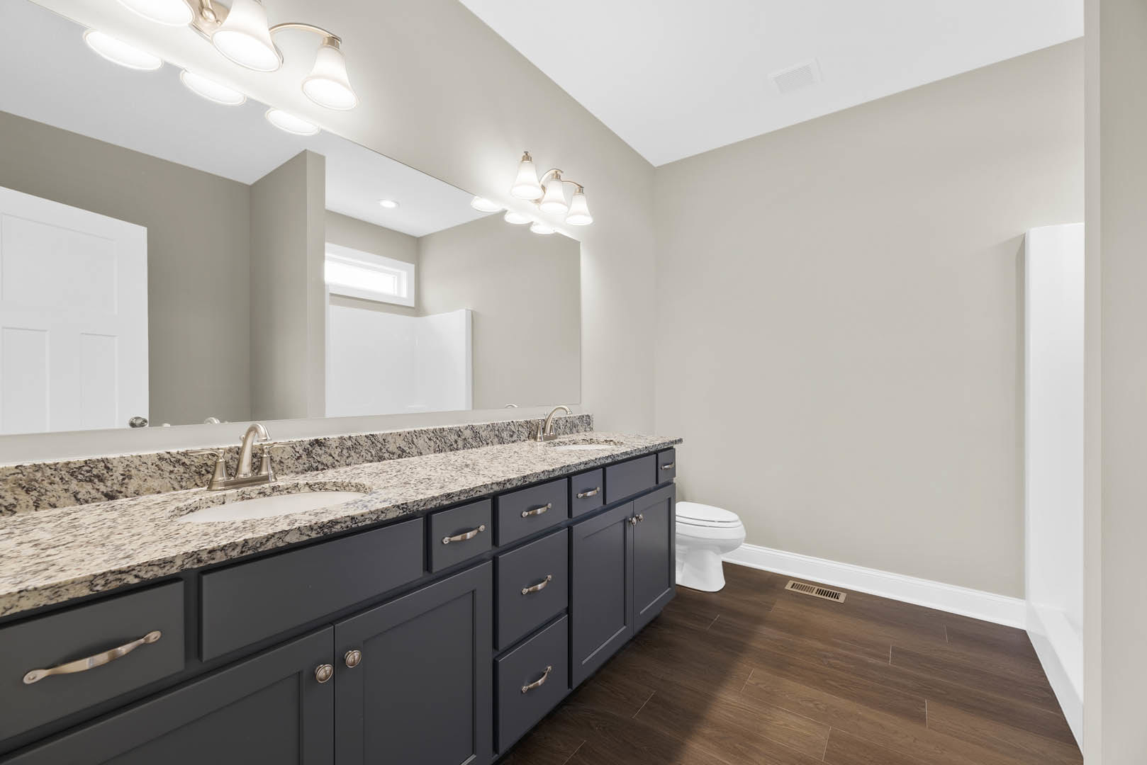 Bathroom with a wide frameless mirror above a rectangular sink, chrome faucet, white countertop, light fixture with multiple bulbs, white toilet, and tiled walls.