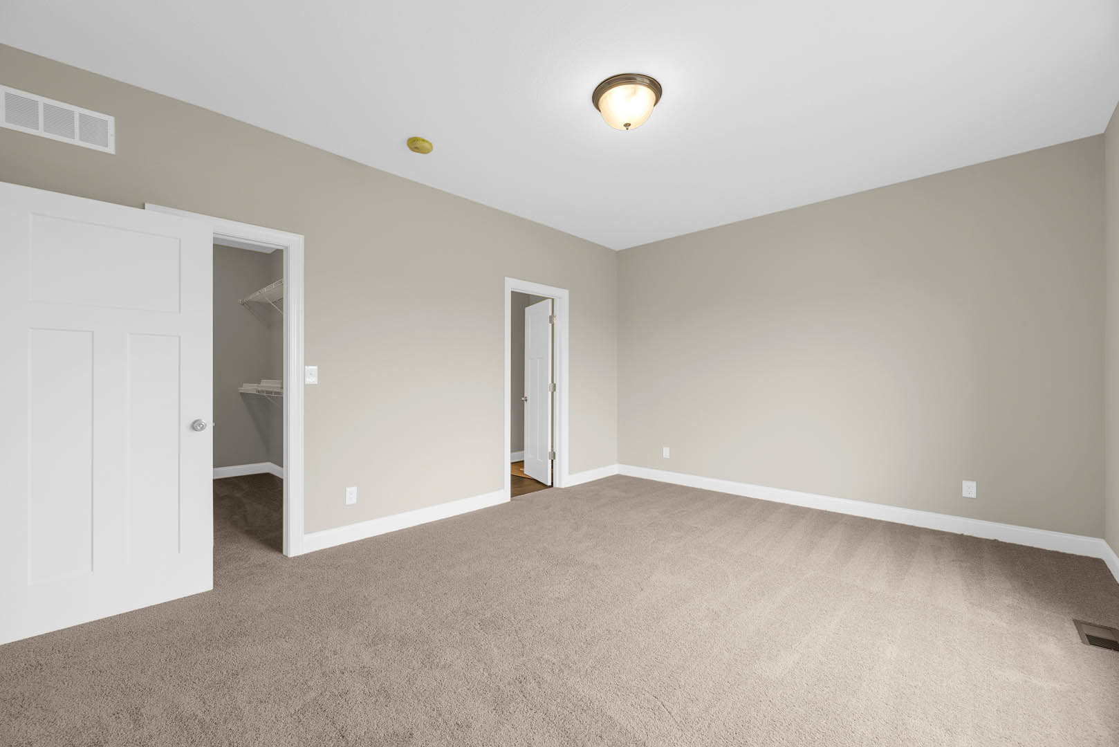 Beige carpeted room with white door featuring silver knobs, ceiling-mounted light fixture, wall vent, and white baseboard molding