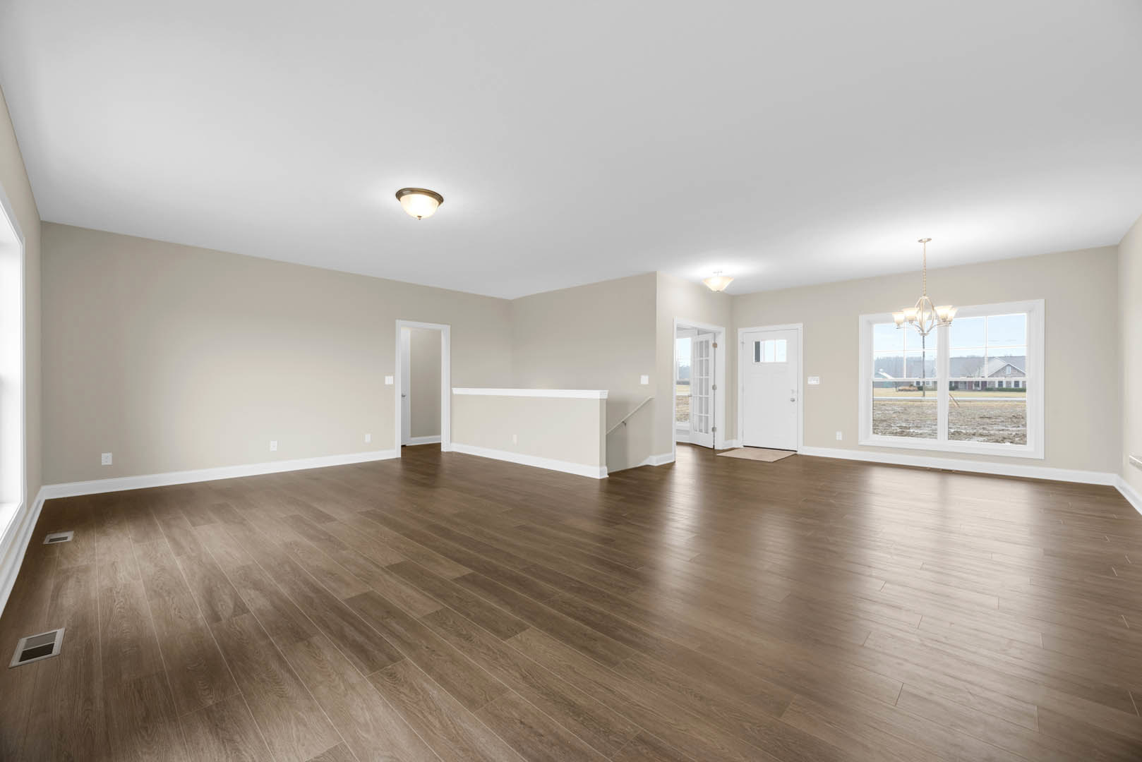 Spacious room with polished hardwood floors, white walls, ceiling chandelier, white door with glass window, wall vent, and large window overlooking grassy field