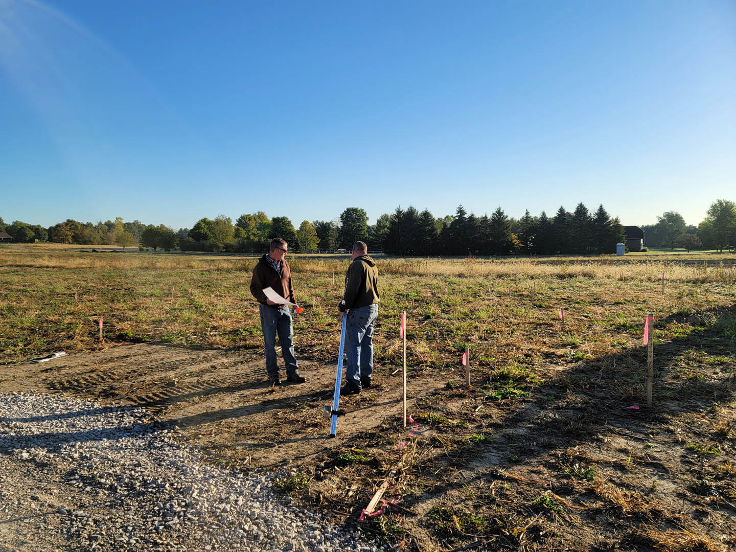 Two men standing in a grassy field with trees and blue sky in the background, one holding a piece of paper and a bottle, the other holding a ruler
