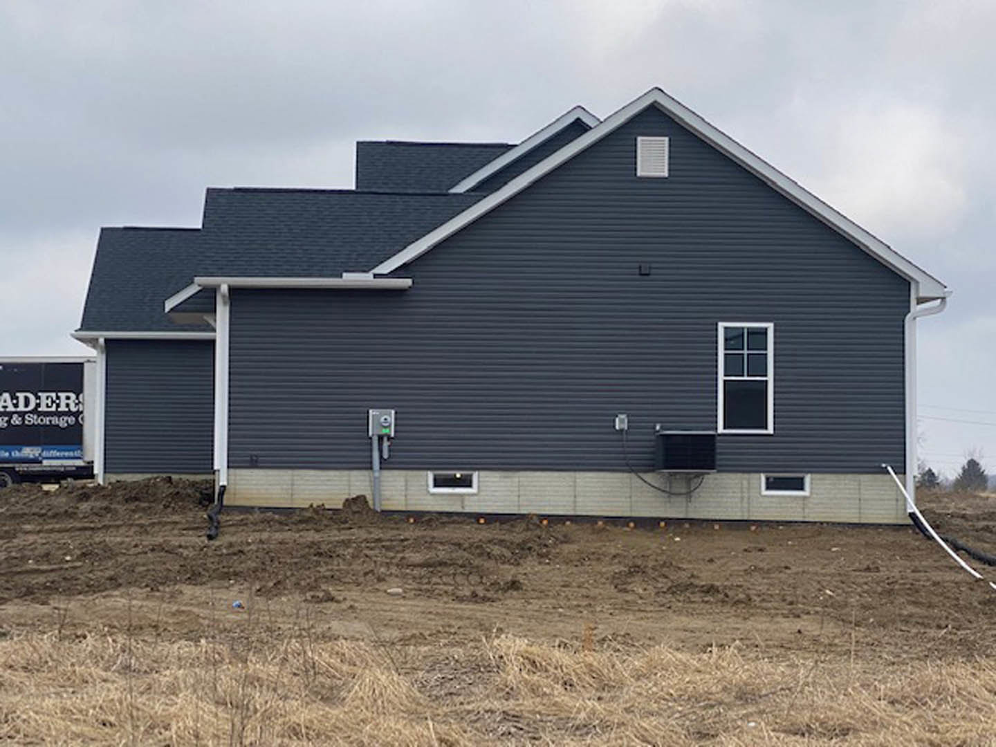 Modern cottage-style home with white siding, black trim, and large windows, set beside a dirt field under a partly cloudy sky