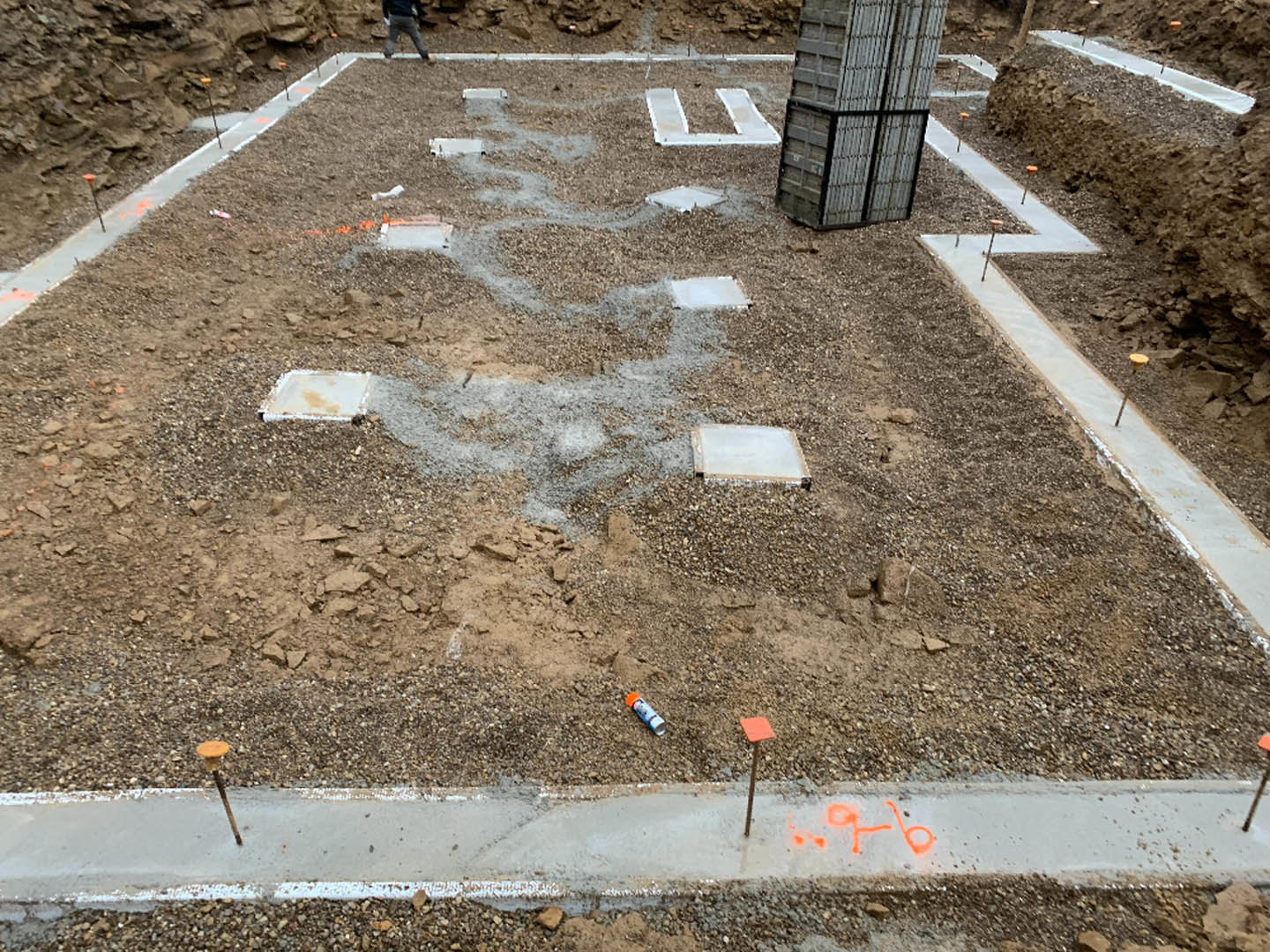 Construction site with exposed soil, stacked concrete slabs, scattered building materials, and a person walking near a white square object on the ground