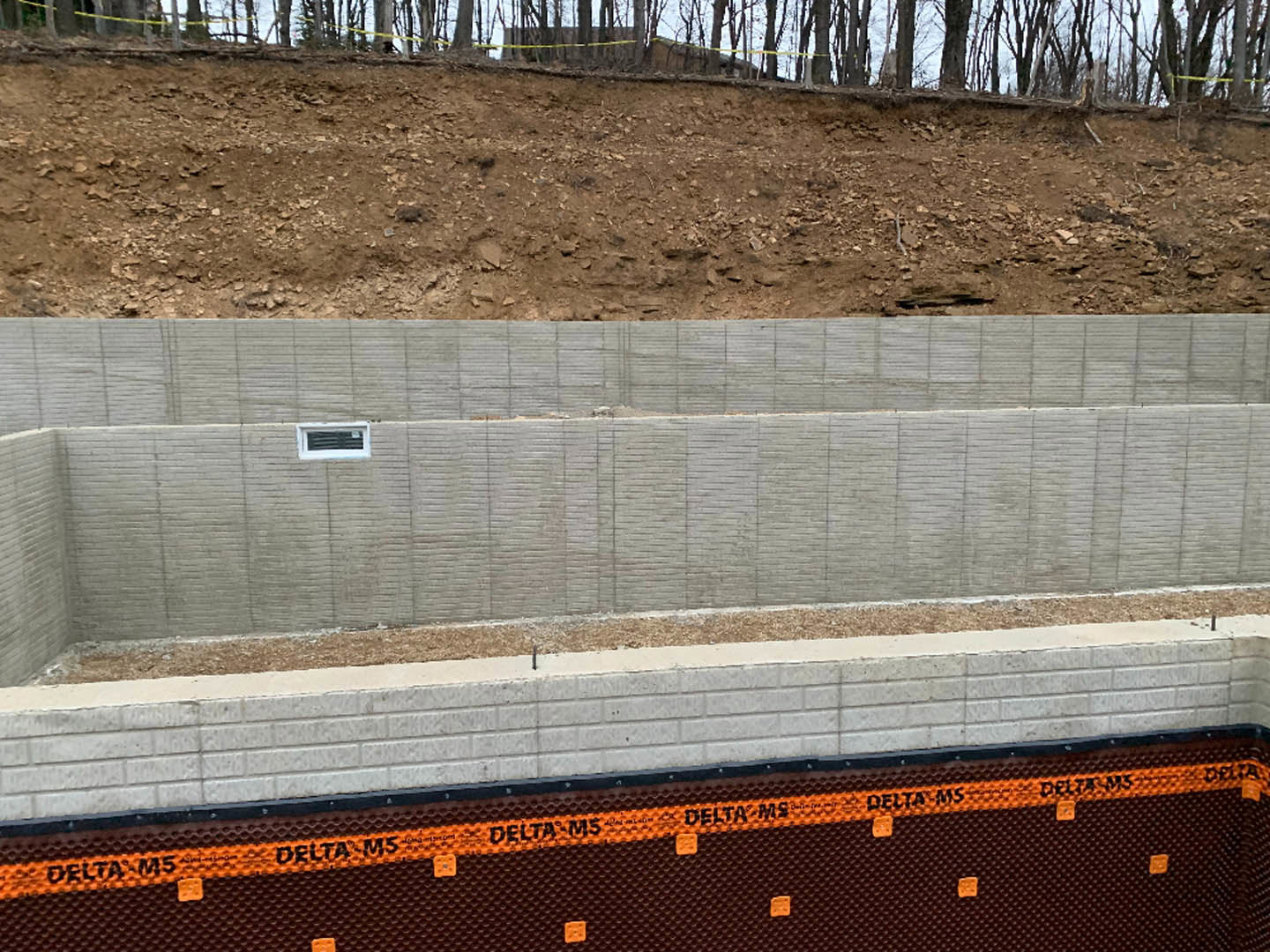 Composite exterior wall with vent, dirt hill bordered by yellow caution tape, partial view of fence and tree in outdoor setting