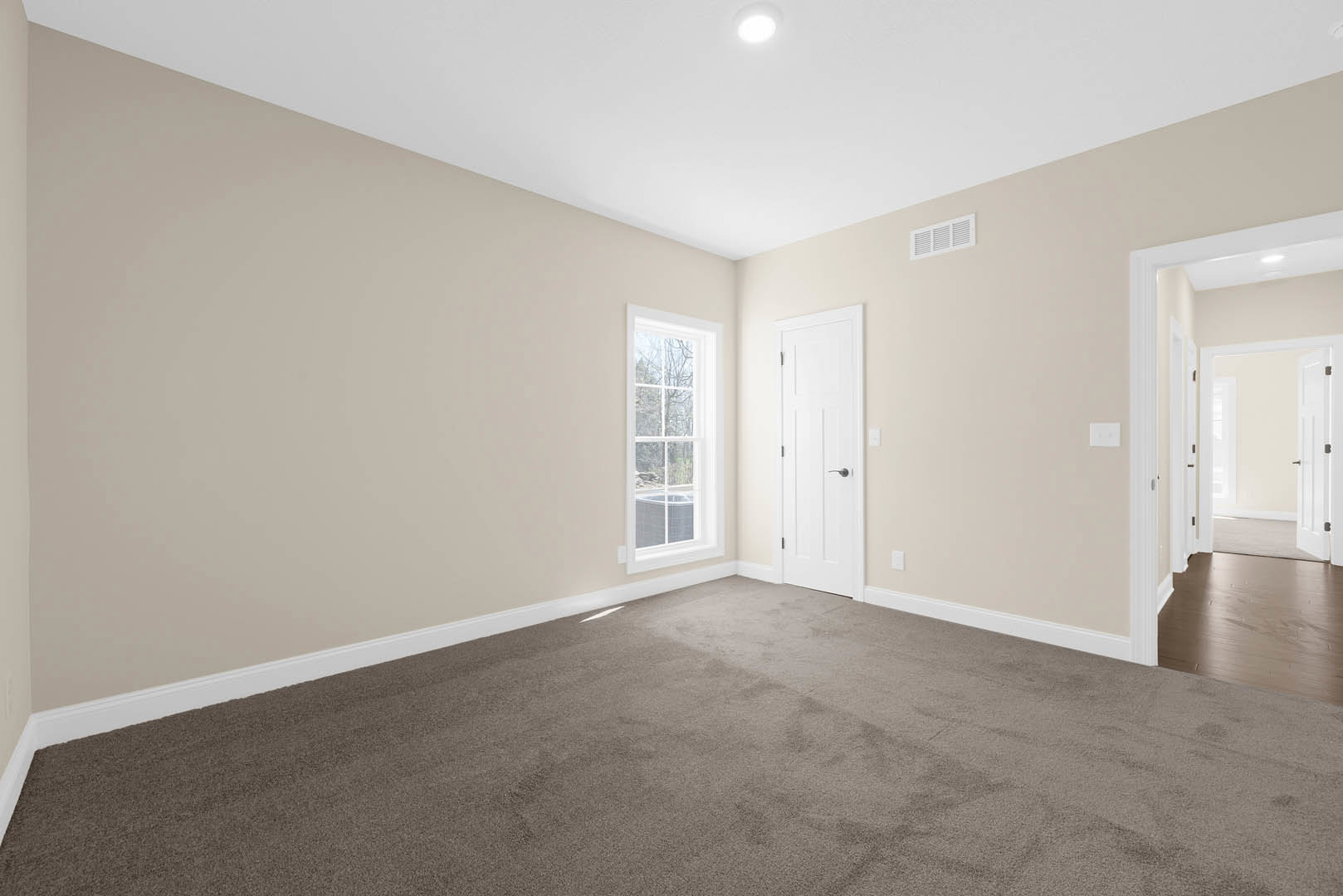 White-walled room with plush beige carpet, white door featuring a silver handle, large window overlooking forested landscape, ceiling light fixture, wall vent, and simple base