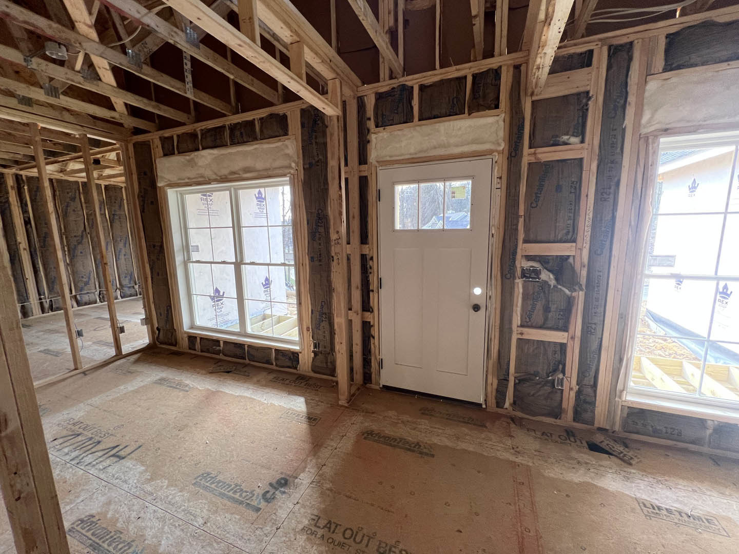 Unfinished room with exposed wooden ceiling beams, white door with glass panel, large window covered in protective plastic, and construction materials visible