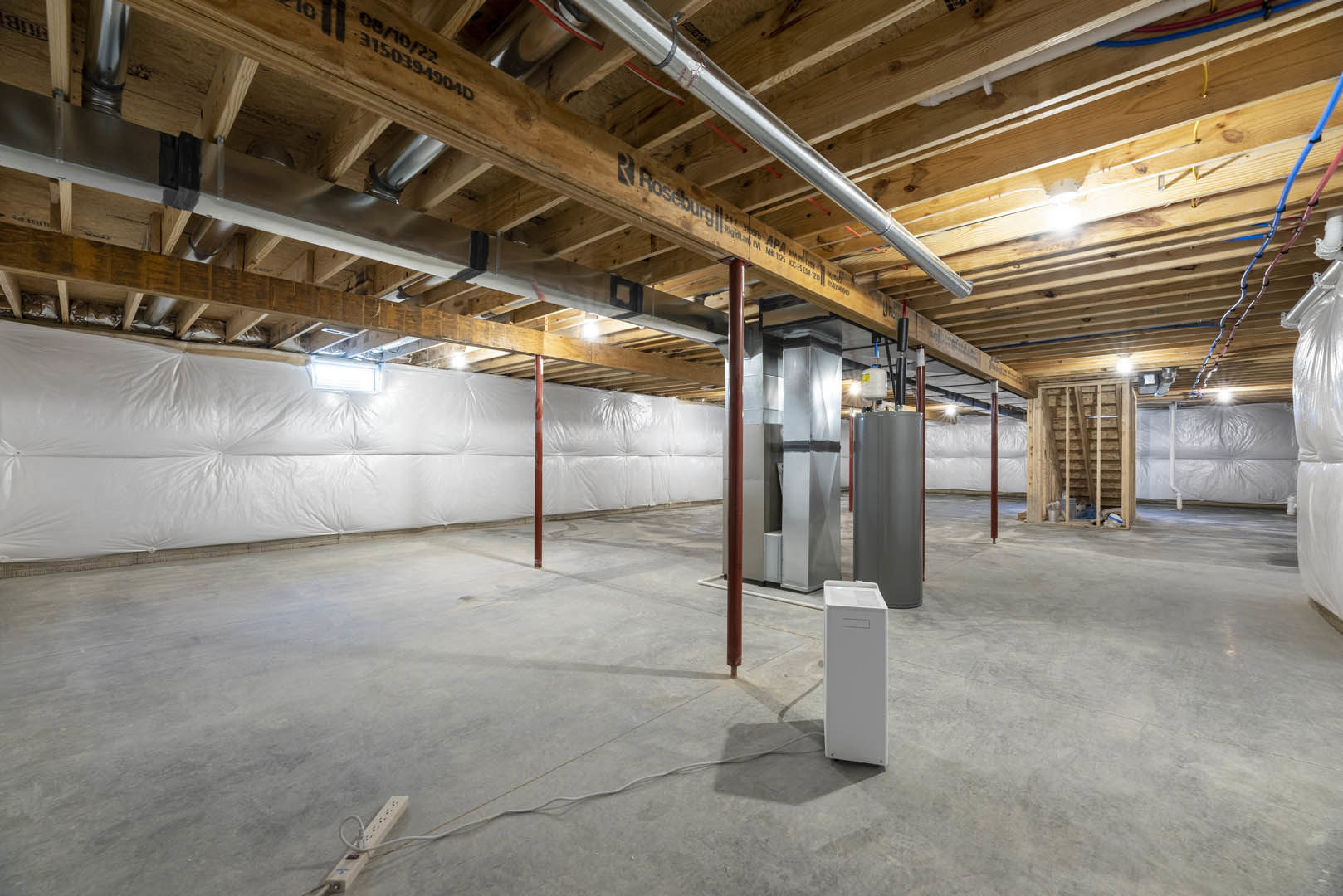 Basement room with exposed steel beams and metal pipes on a wooden ceiling, white wall partially covered in plastic, white rectangular utility boxes on concrete floor.