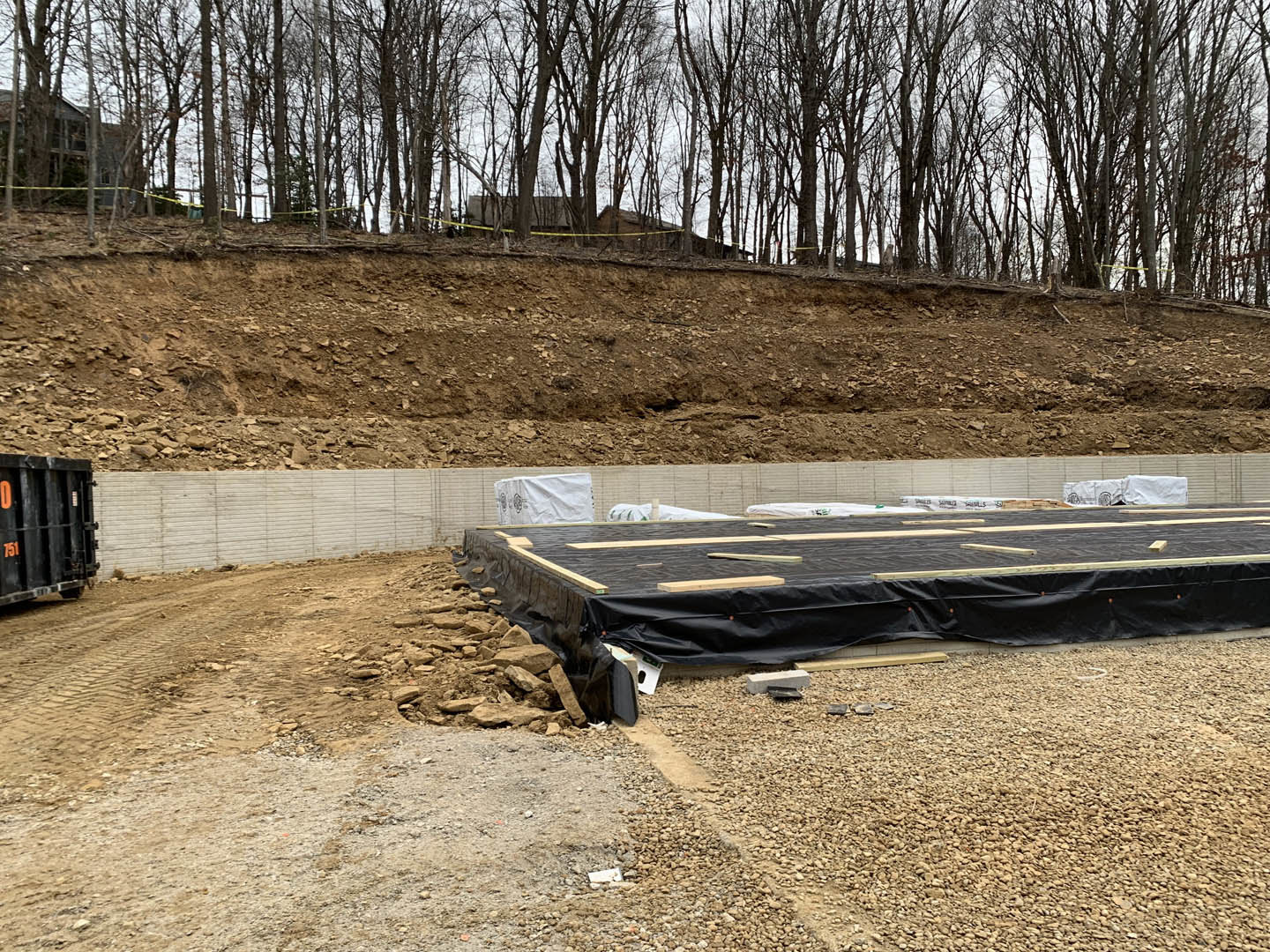 Black tarp covering foundation at wooded construction site with exposed soil and scattered building materials