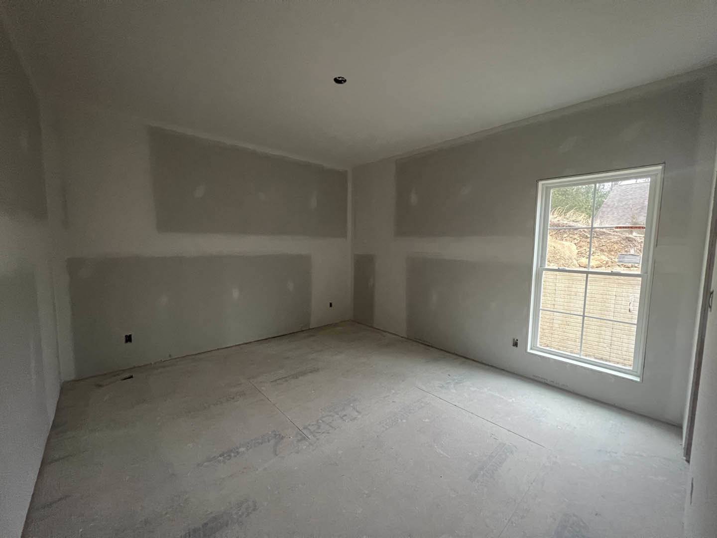 Room with white plaster walls and ceiling, concrete floor, large window overlooking dirt field, person standing near window, black ceiling fixture visible