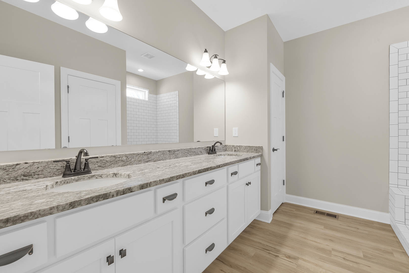 Bathroom with expansive mirror above white cabinetry, row of lights mounted over sink, chrome faucet, white paneled door with hinge, wood flooring featuring vent near baseboard