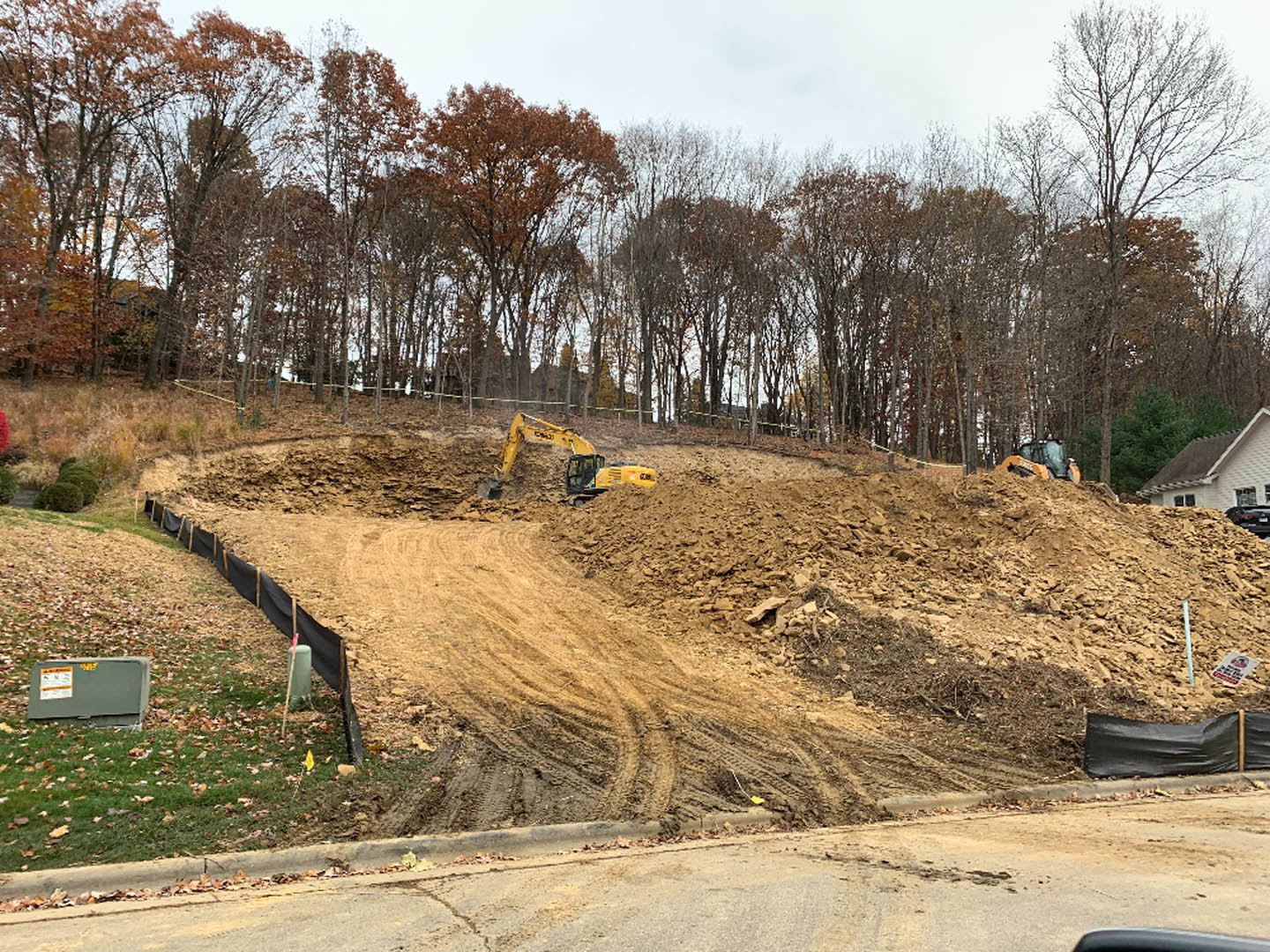 Bulldozer excavating a dirt hill on a residential construction site, surrounded by trees and fenced area with green utility cylinder