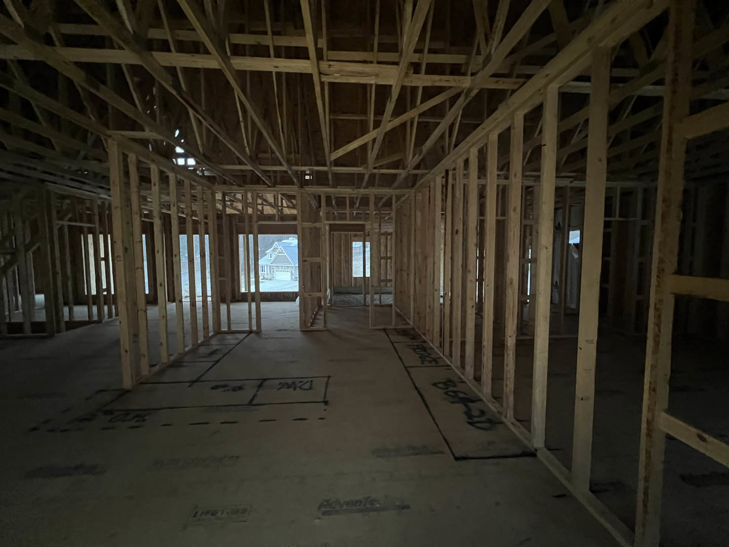 Wood-framed house under construction with exposed ceiling beams, subfloor marked with black writing, visible garage area, and close-up of a logo on building materials.