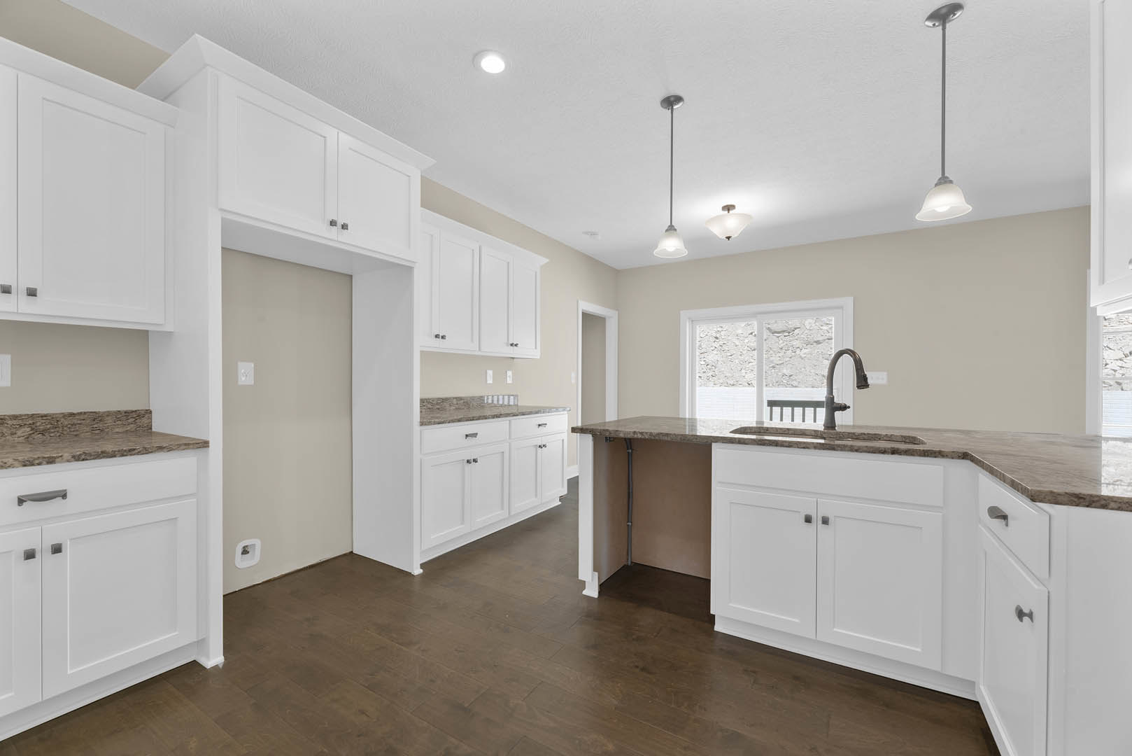 White kitchen cabinets with brushed metal handles, dark brown wood flooring, white-framed door, large window with natural light, stainless steel sink and appliances, woman standing
