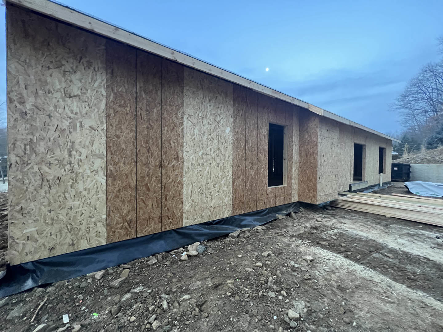 Wood-framed custom home under construction with exposed beams, blue tarp covering materials, dirt ground with scattered rocks, and clear blue sky overhead