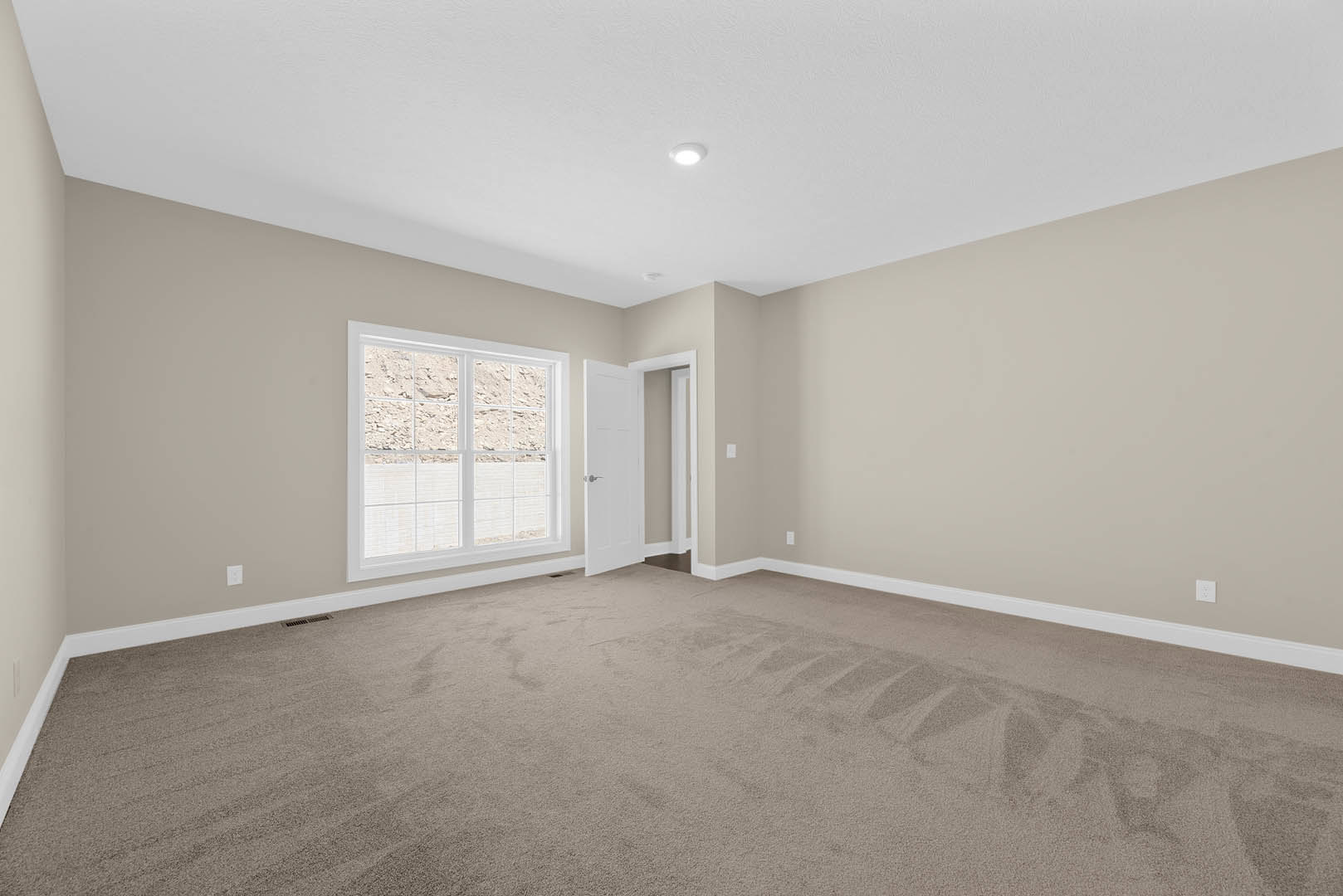 Carpeted room with white door featuring silver handle, window overlooking rocky landscape, ceiling light fixture, white walls with molding