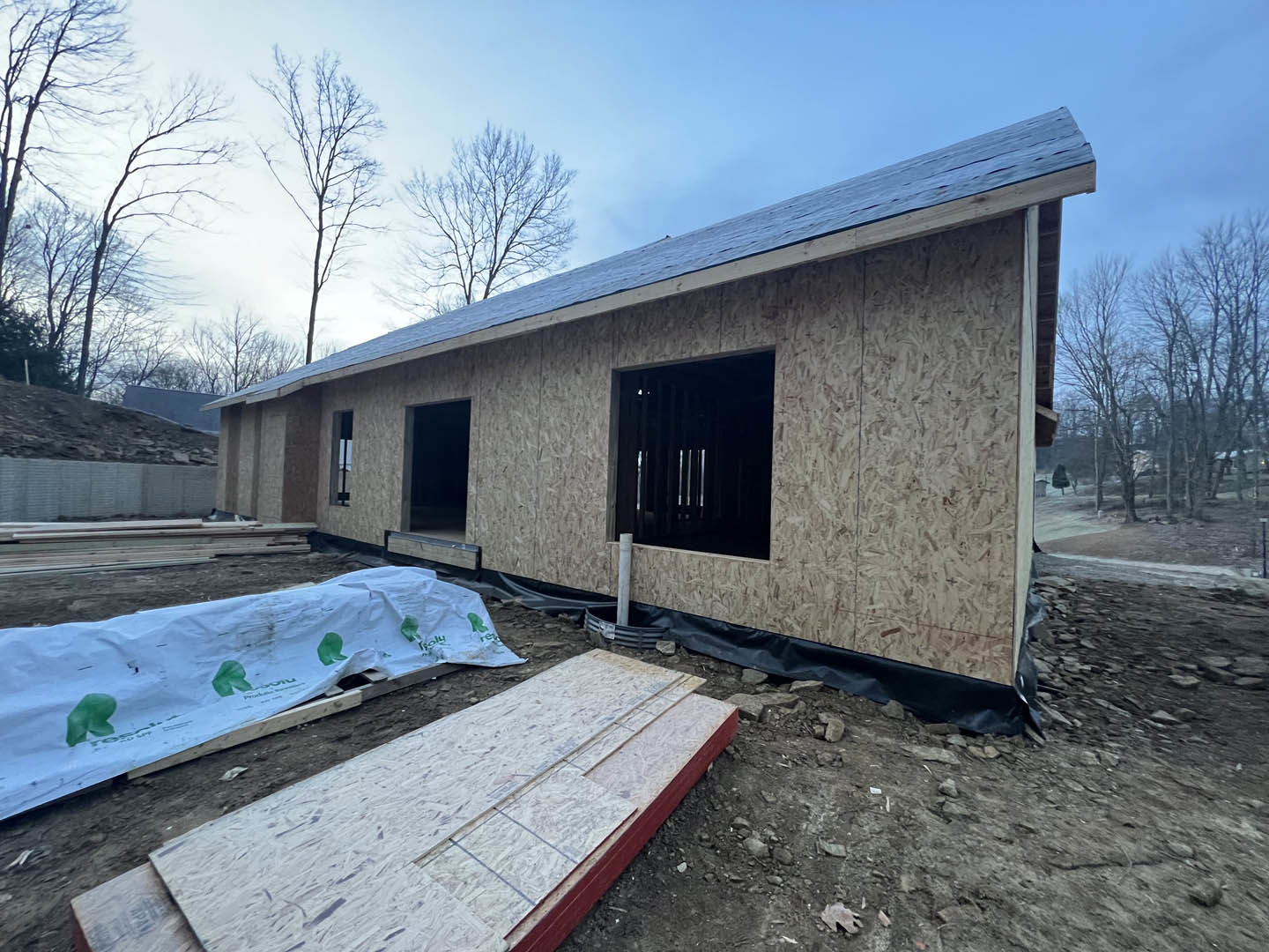 Partially built house with exposed plywood, barred door, white tarp featuring green logo draped over construction area, scattered building materials on ground, tree branches