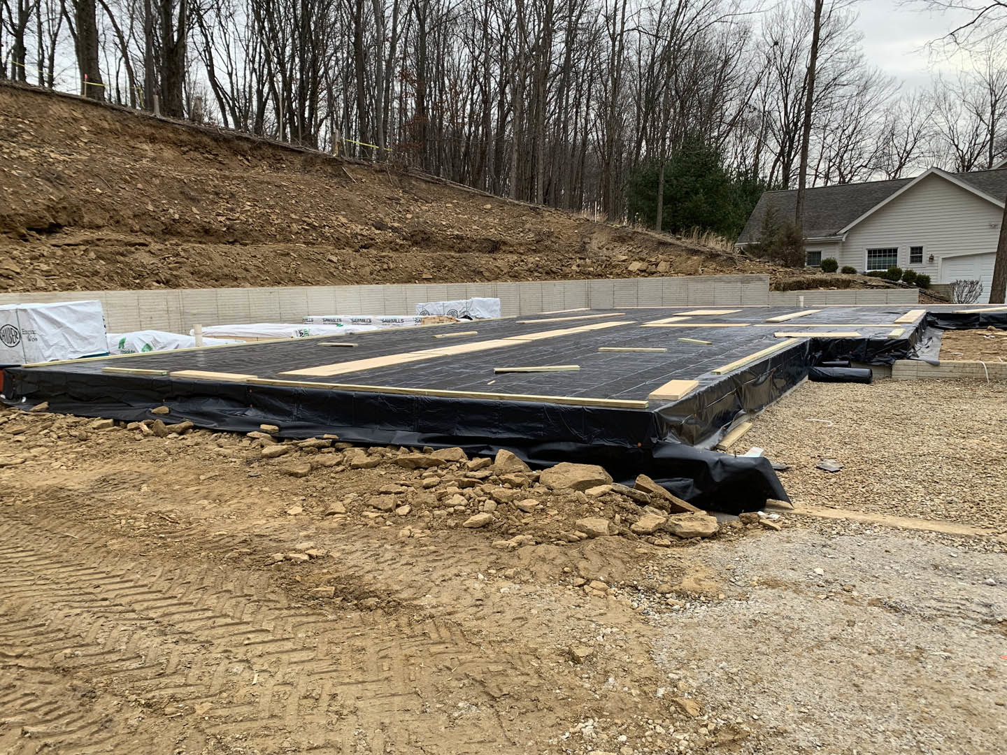 Black tarp covering dirt ground at residential construction site, white sheet with black writing nearby, bare trees and house in background.
