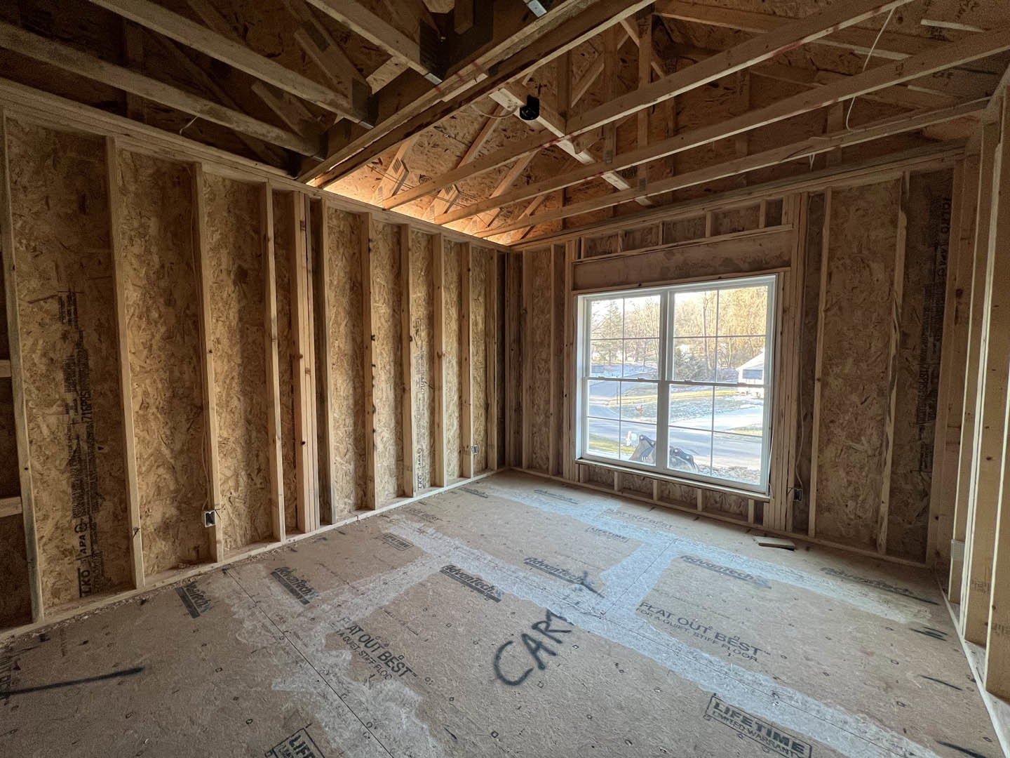 Living space with two large windows, exposed wooden ceiling beams, light wood flooring, and view of road and trees outside.