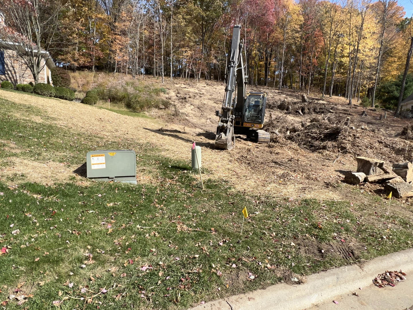 Framed custom home under construction with exposed wood beams, crane lifting materials, dirt lot surrounded by trees, yellow warning signs on equipment, bulldozer parked near grey