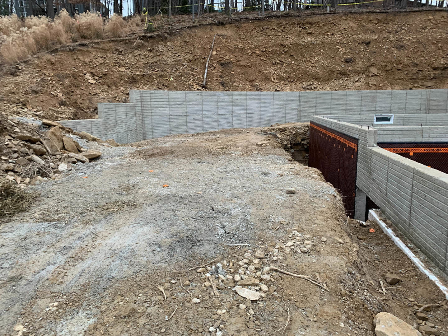 Excavated foundation with exposed concrete wall, surrounding soil, and patches of tall grass adjacent to a wooden fence