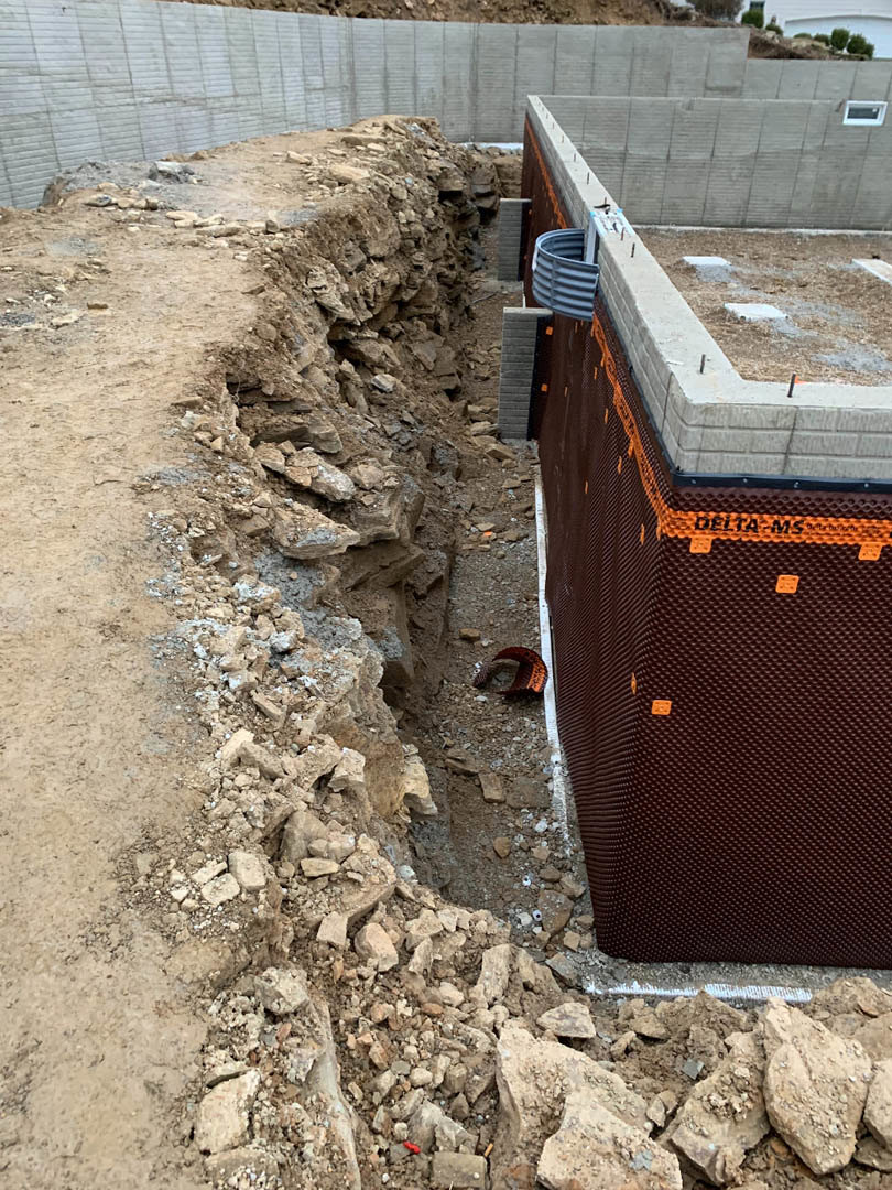 Concrete exterior wall with exposed hole in the ground, grey plastic pipe visible among dirt and rocks