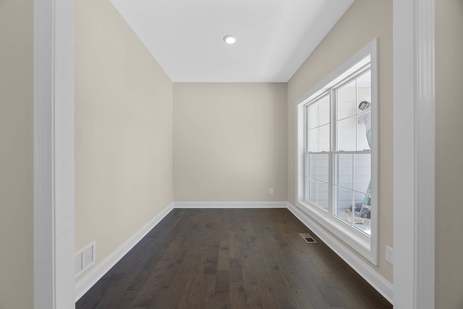 Dark hardwood floor with white baseboard trim, large window, ceiling light fixture, painter working near window, close-up of floor vent