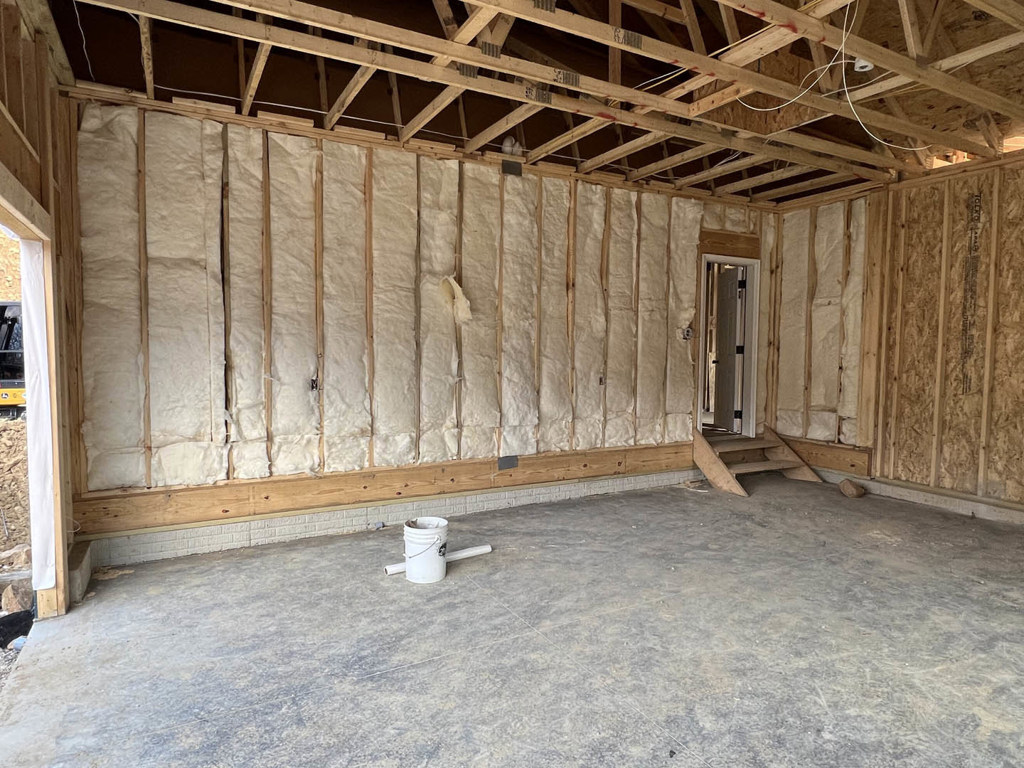 Unfinished room with exposed wooden ceiling beams, concrete floor, white bucket near wall, and partially installed insulation