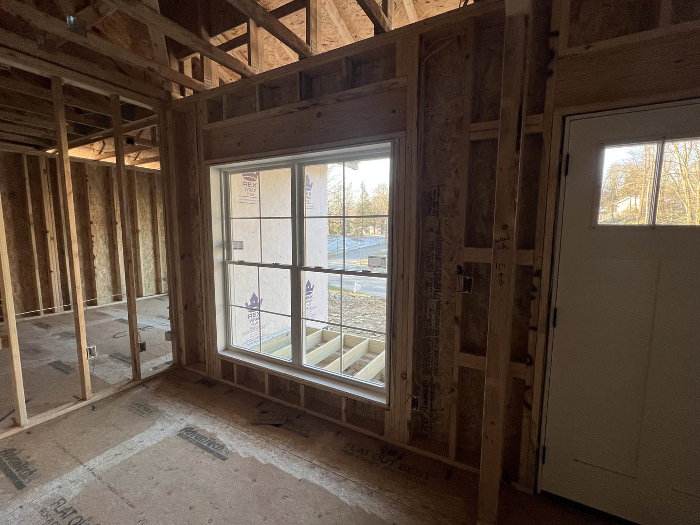 Sunlit room featuring a wood-framed window with views of trees, white door with glass insert, light hardwood flooring, and exposed ceiling beam
