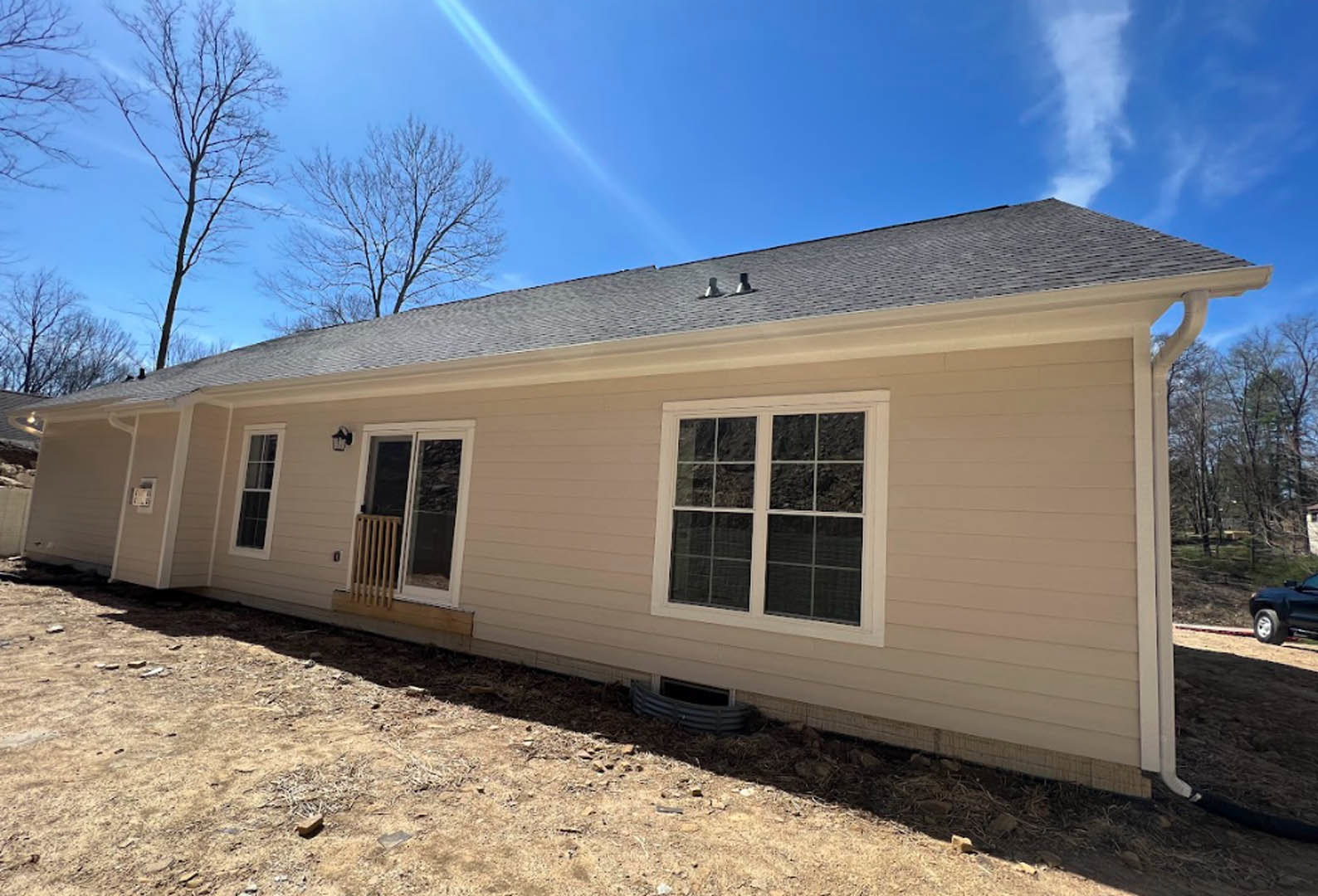 Framed house under construction with exposed wood siding, surrounded by green trees and under a clear blue sky