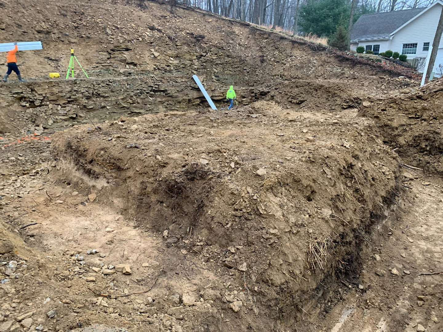 Dirt hill with two people, one in an orange shirt, carrying material; house partially visible behind hill, surrounded by trees and plants