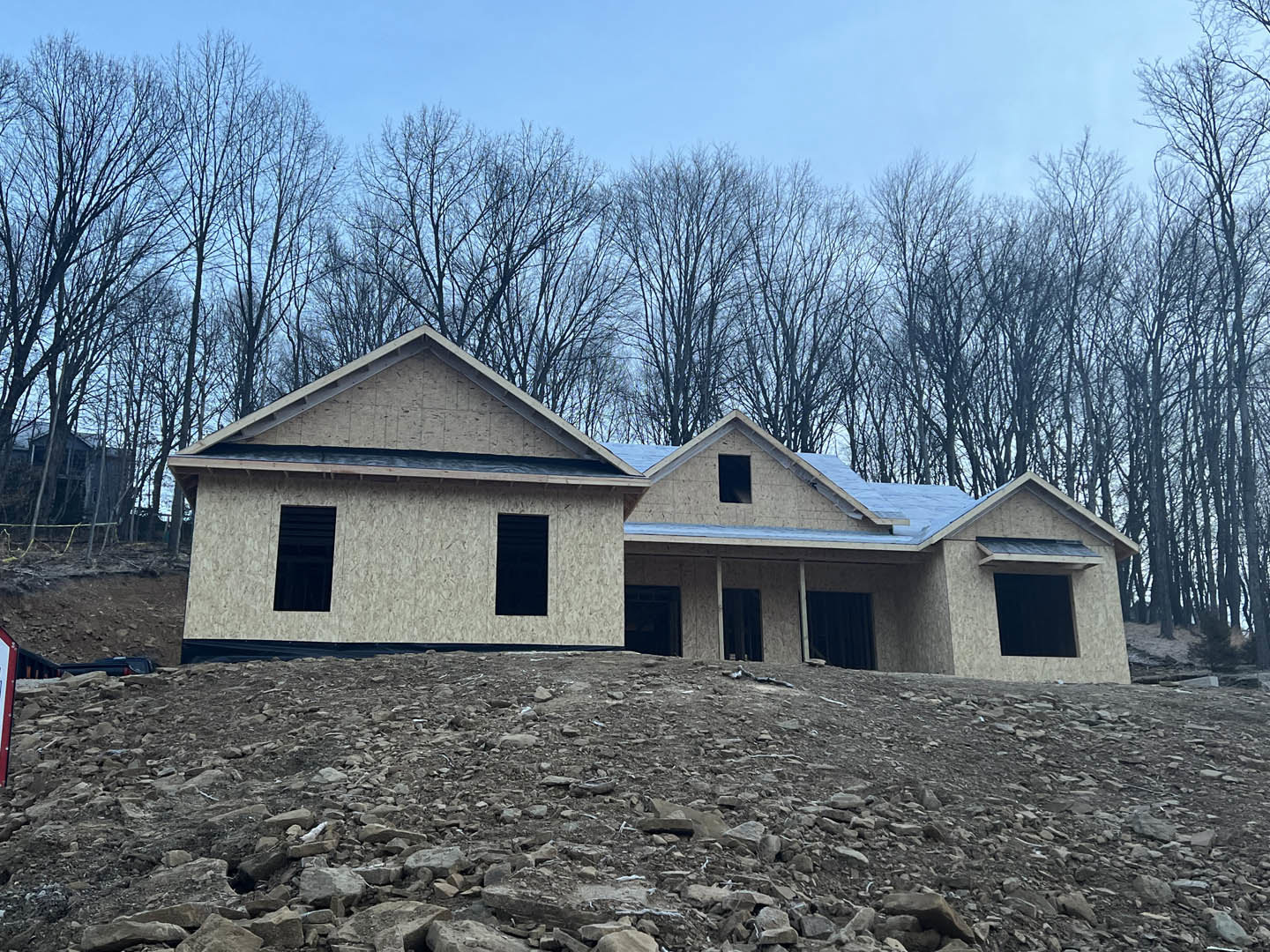 Modern two-story home with light siding and large windows, situated atop a rocky hill, surrounded by mature trees and a clear sky in the background
