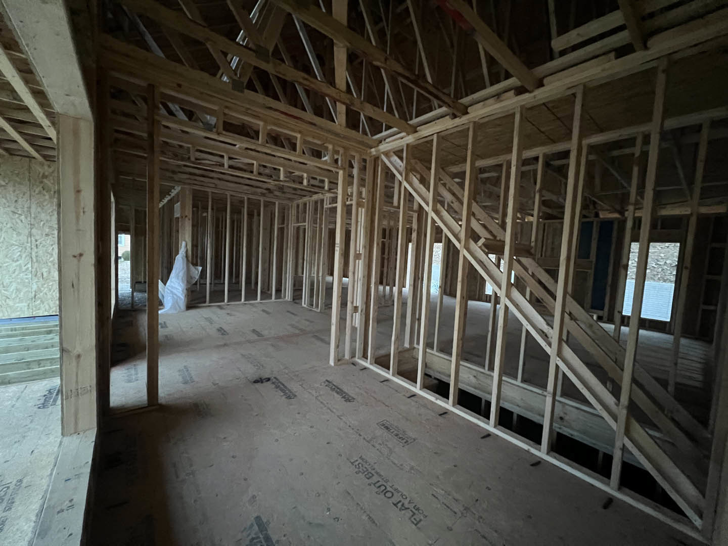 Exposed wooden framing and beams inside a partially constructed house, with a white insulation bag hanging from a post, unfinished grey subfloor, and a window overlooking a rocky