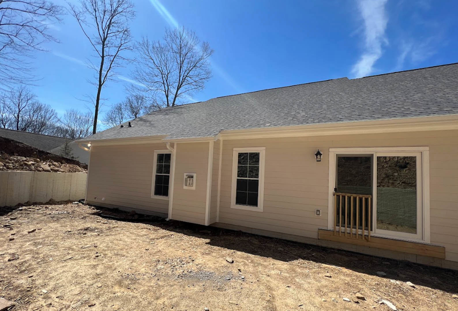 Two-story home with gray siding, white-framed windows, covered porch, sliding glass door with wooden railing, landscaped yard, mature trees, and partly cloudy sky in the