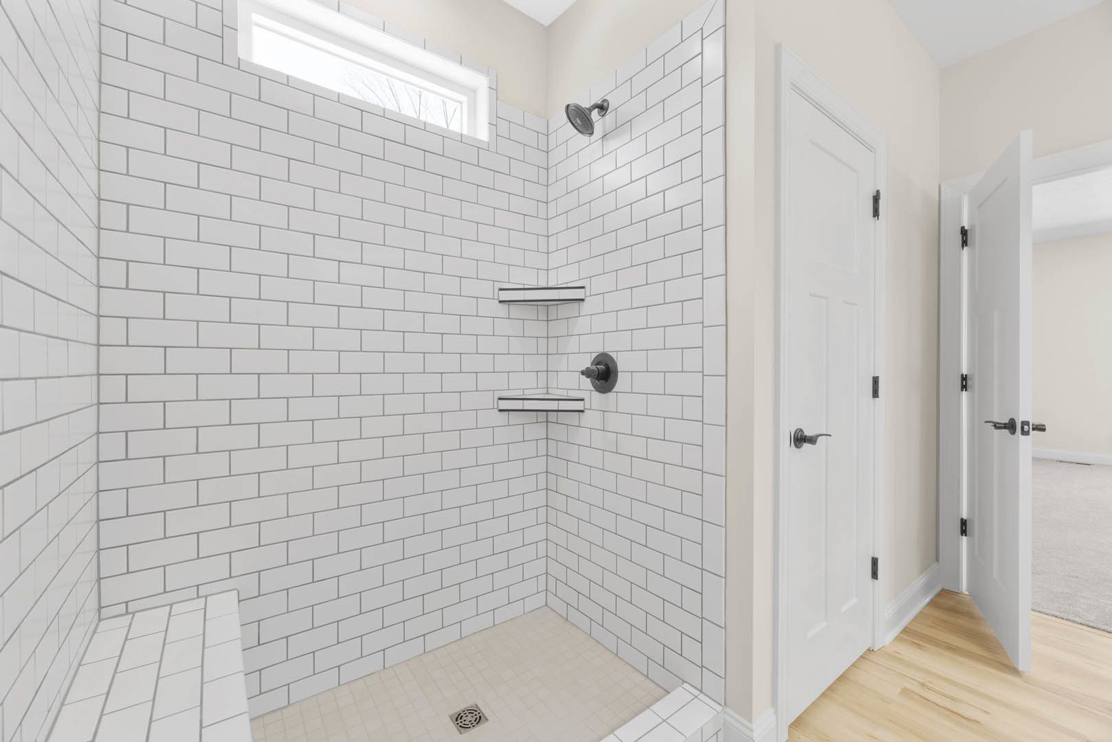 White tiled shower with two chrome shower heads, white brick accent wall, frosted window, metal drain on tiled floor, partial view of white door.