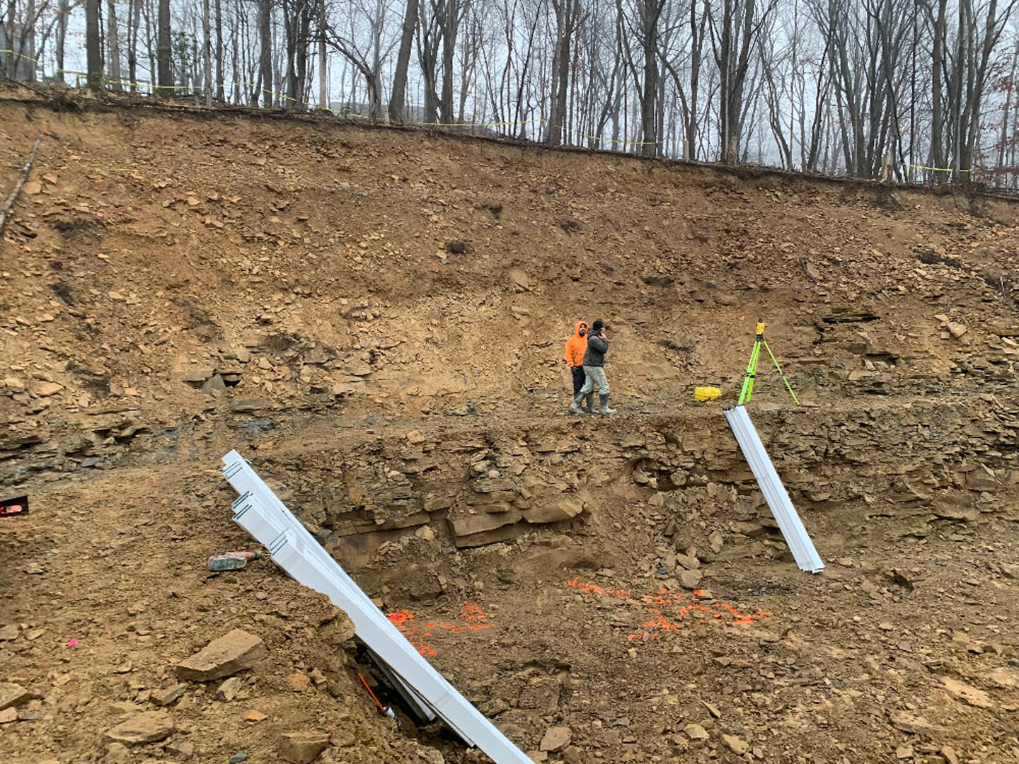 Group of people walking across a dirt hill with scattered white metal beams and orange sticks, surrounded by trees in the background