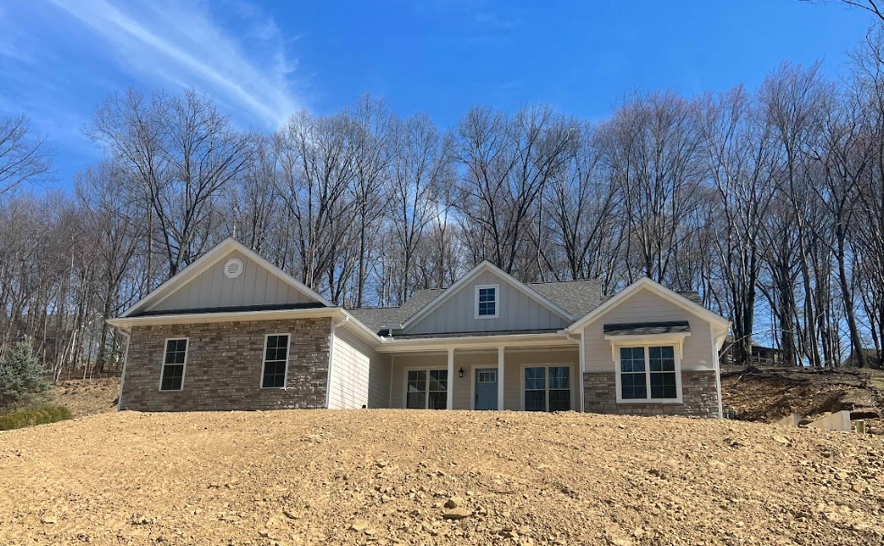 Two-story home with blue front door, brick chimney, multi-pane windows, and a large dirt hill in the foreground; trees and clouds in the background.