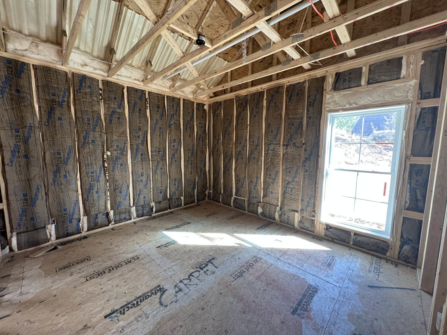 Room under construction with exposed wood walls, ceiling beams, white-framed window, floor insulation, and visible roof truss