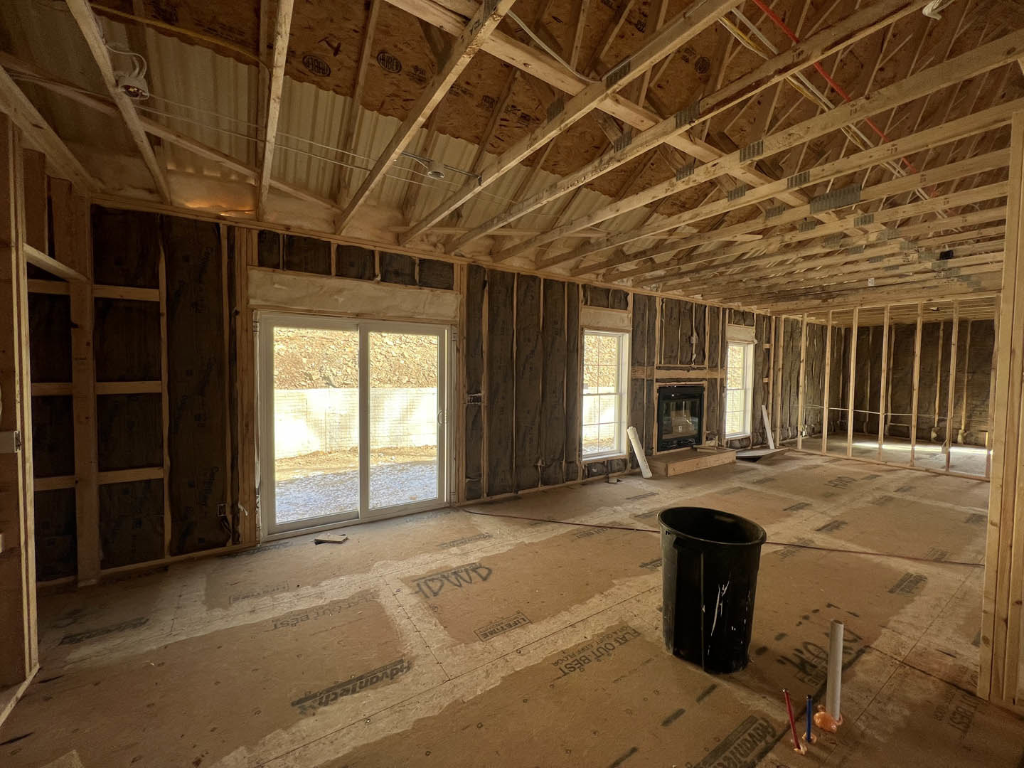 Living room under construction with exposed wooden ceiling beams, sliding glass door, fireplace, cracked black trash can, and unfinished floors.