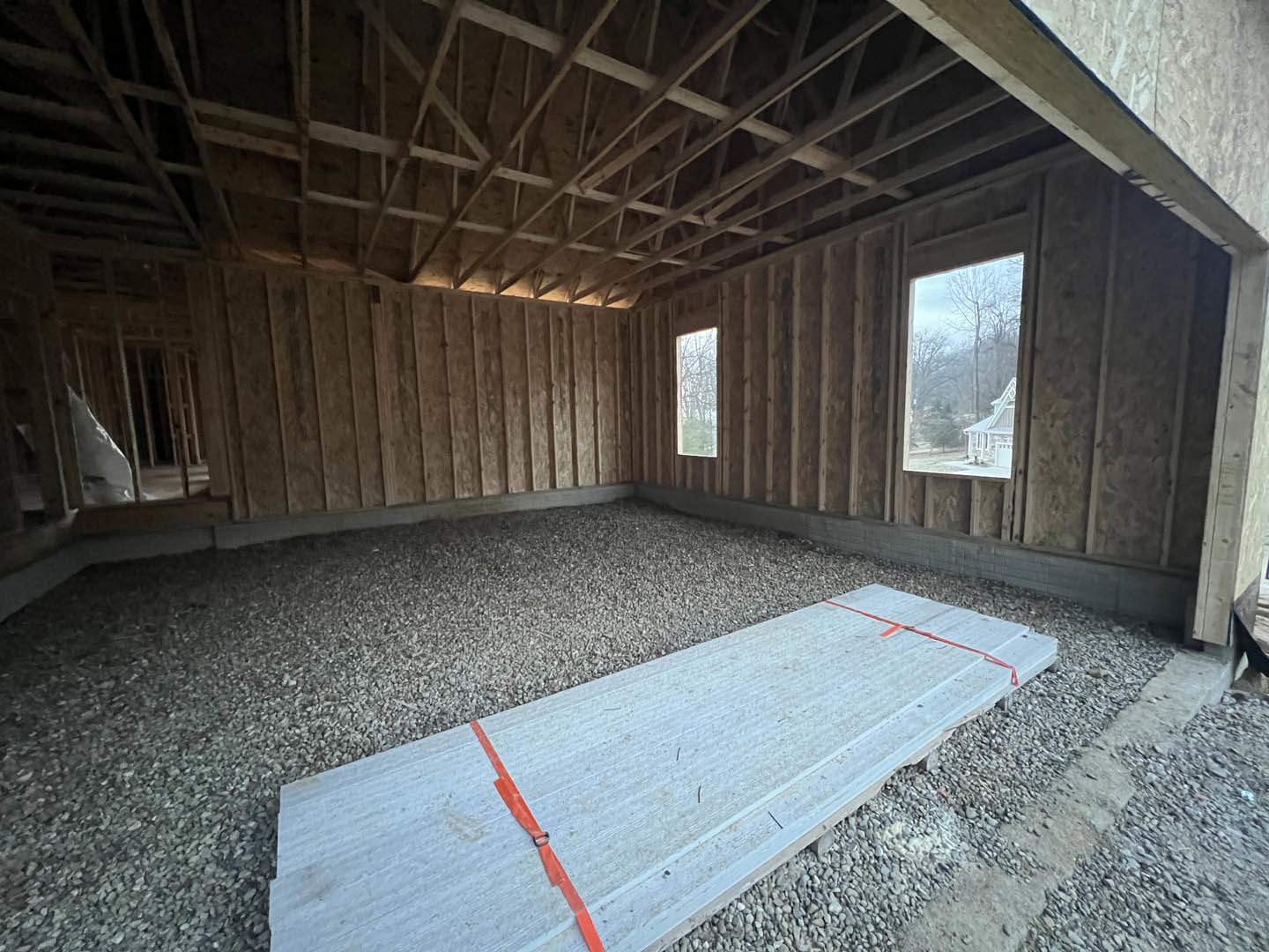 Wide wooden plank with red tape resting on concrete floor, exposed wooden ceiling beams, large window revealing leafy trees outside, unfinished walls and building insulation