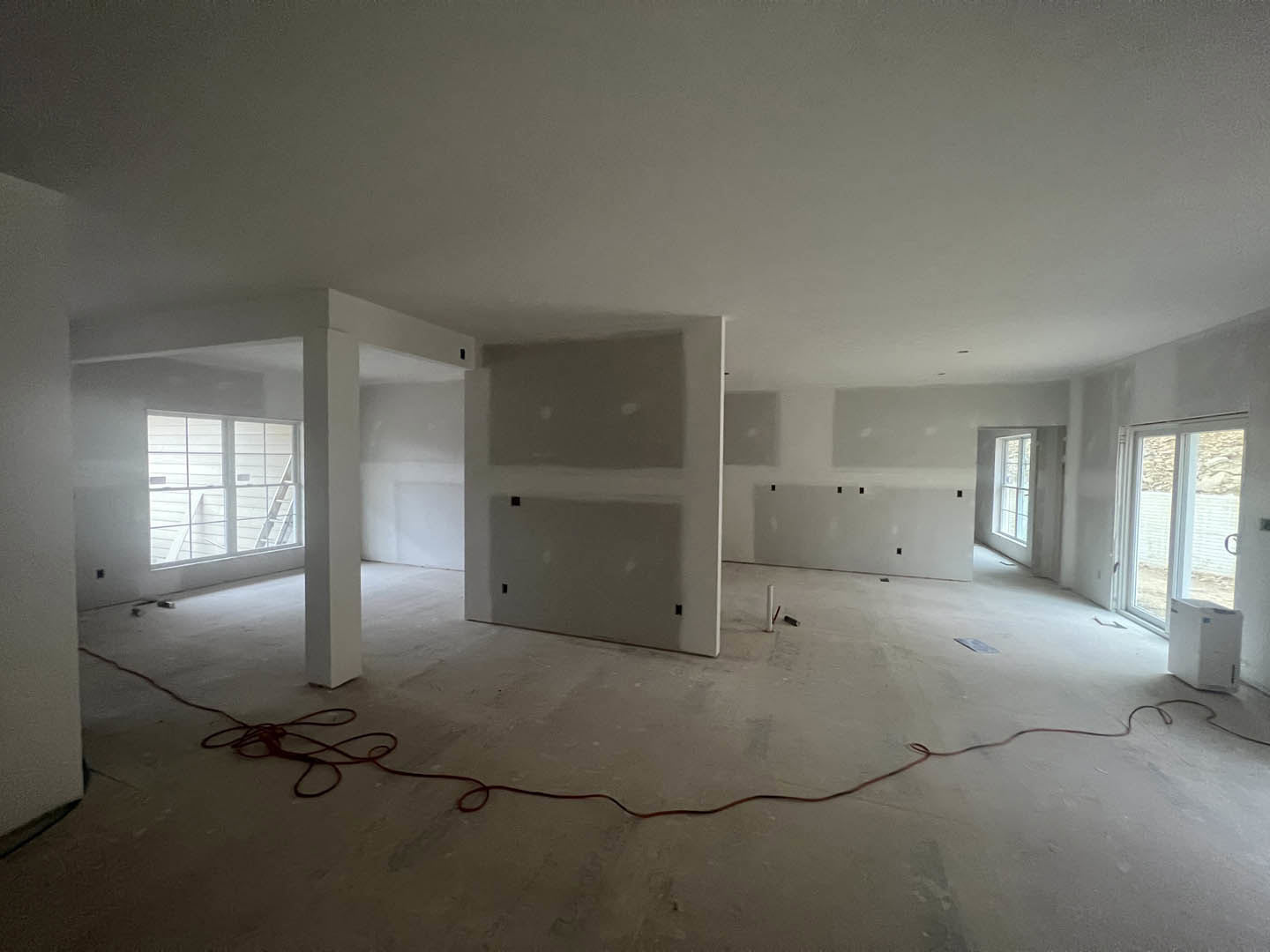 Unfinished room with plaster walls, red electrical wire on the floor, window with ladder in background, white shelf, white box resting on person's lap, close-up of door.
