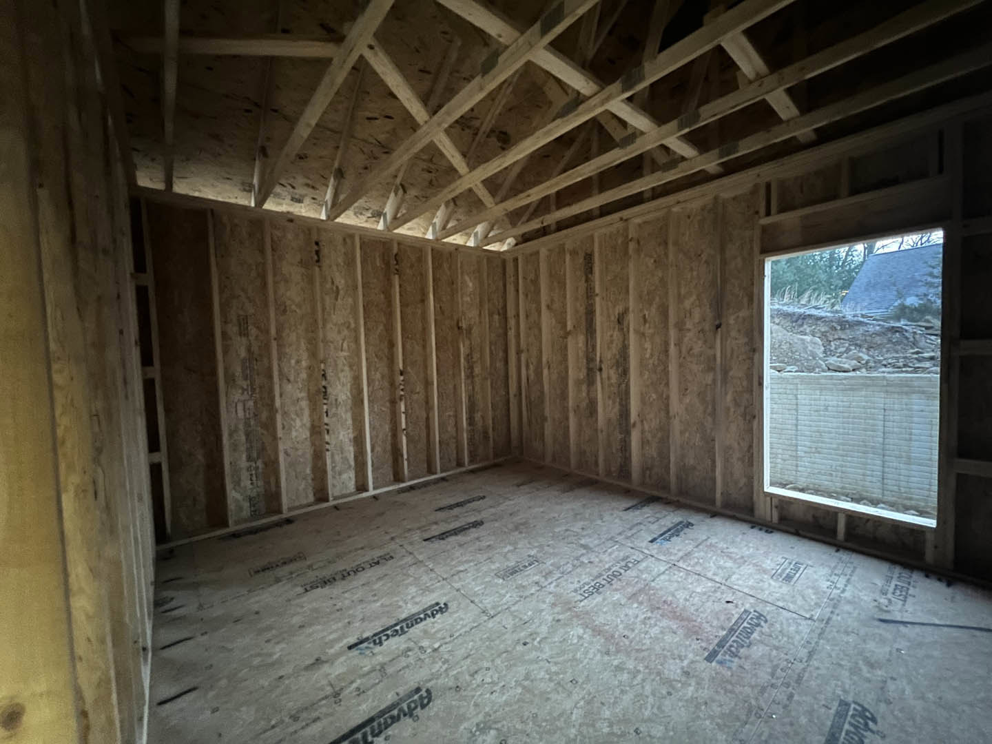 Room with exposed wood ceiling beams, large window overlooking a house and hillside, light-colored walls, and floor featuring black text or lines.