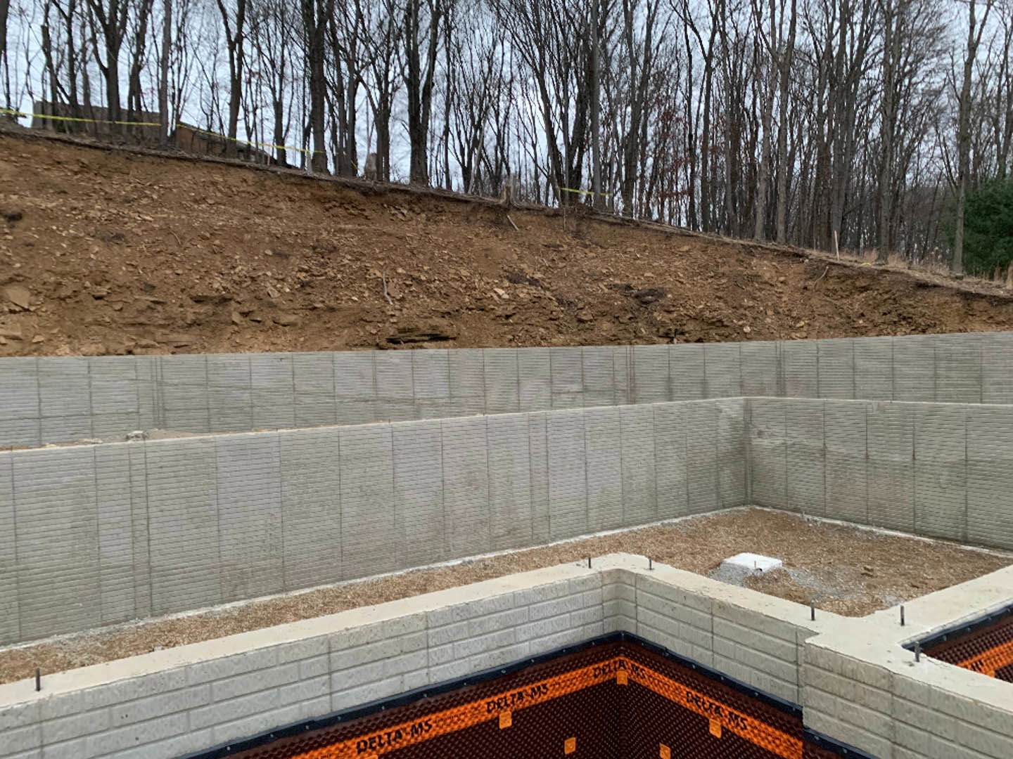 Concrete foundation and retaining wall bordered by grassy hill with mature trees, white utility box in foreground, metal fence along property line, clear sky overhead