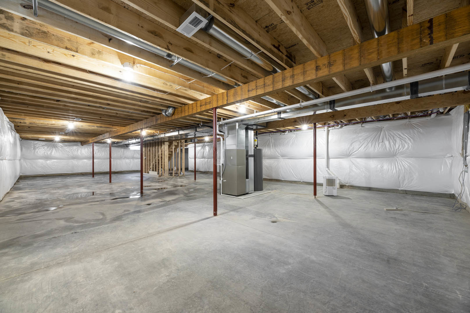 Spacious basement with exposed metal pipes, white vent on ceiling, large white wall, and metal utility box.