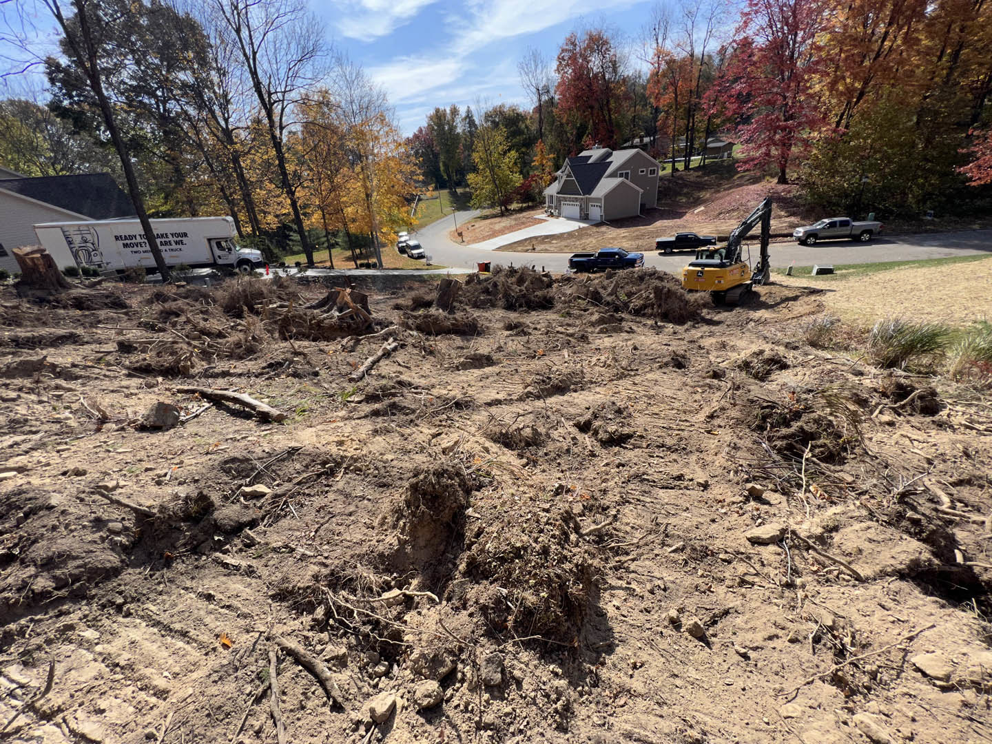 House with attached garage situated on a dirt field, white truck parked nearby, yellow crane and construction equipment in the background, surrounded by trees under a partly cloudy
