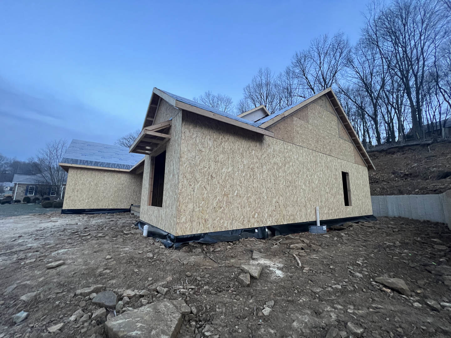 Partially built hillside house with exposed plywood walls, blue metal roof, white framed windows, scattered rocks and construction materials in foreground, trees and blue sky in